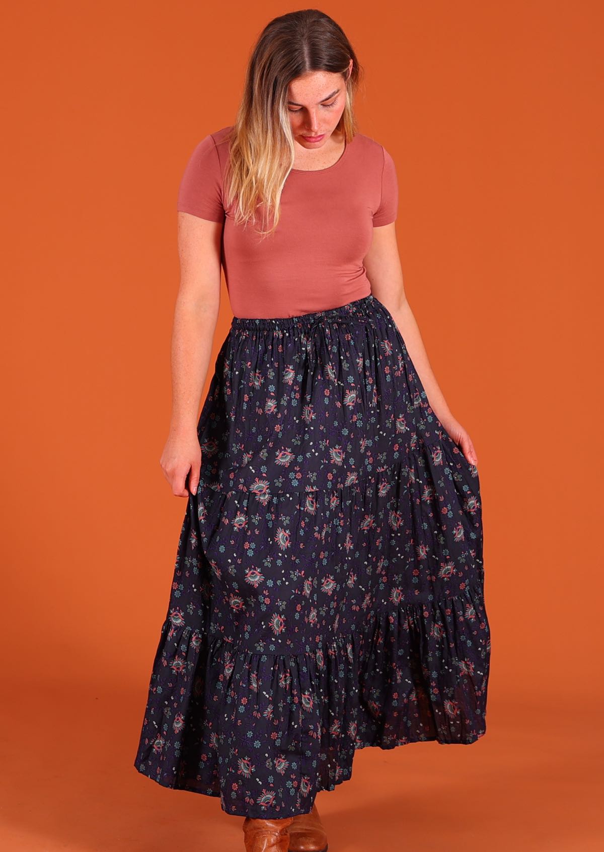 Model standing looking down and holding middle tier of dark Purple 100 percent cotton Indian floral printed three tiered maxi skirt