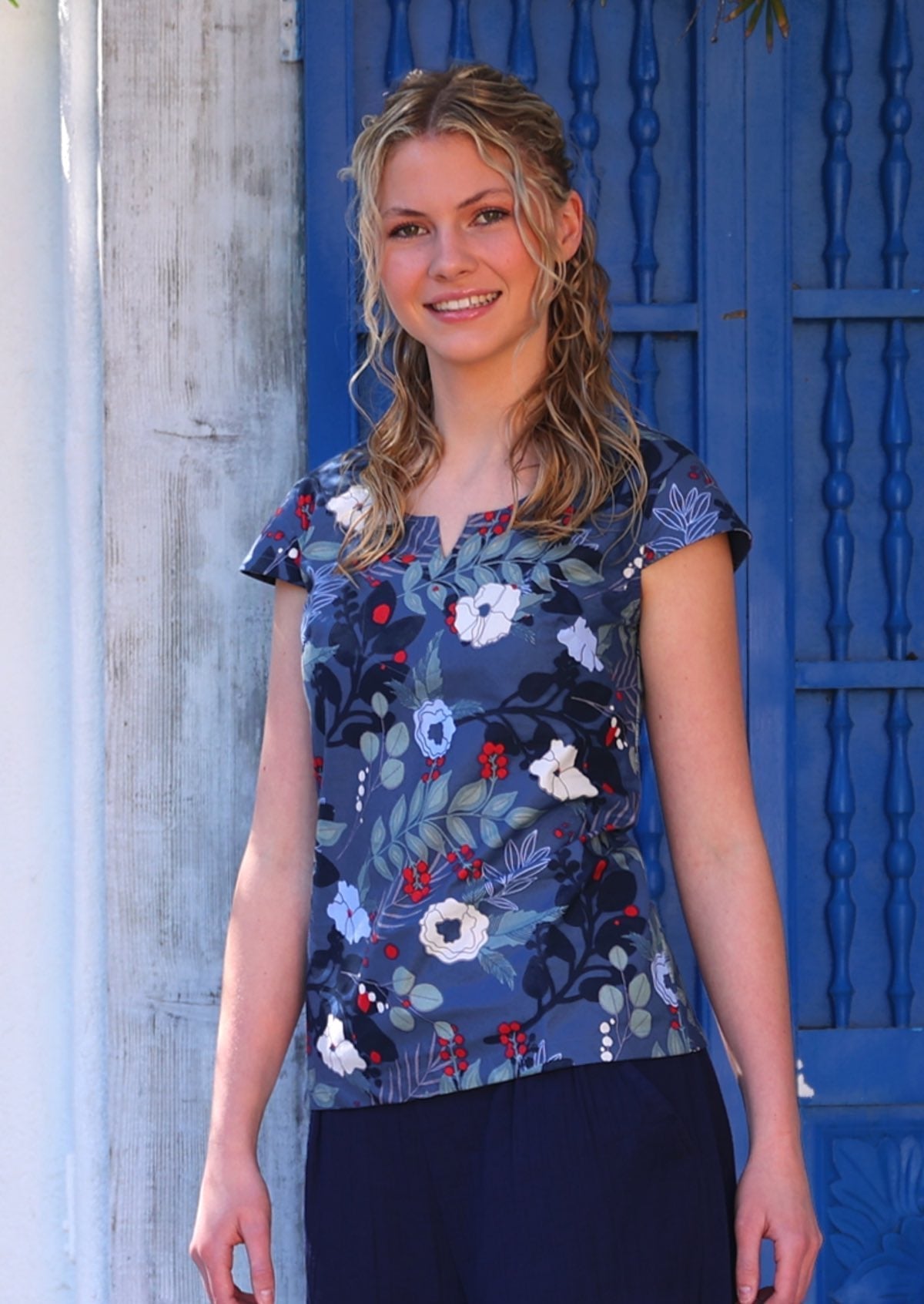 Woman with blonde hair wearing a cotton top with round neckline with keyhole cutout, in blue, white, grey and red floral print, in front of blue door