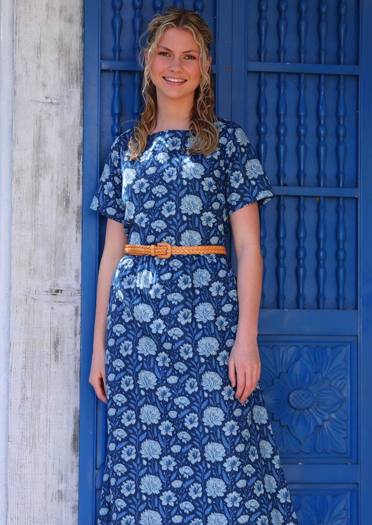 Model standing in front of blue door wearing 100 percent cotton sundress pale flowers on a navy base added a waist belt to define waist