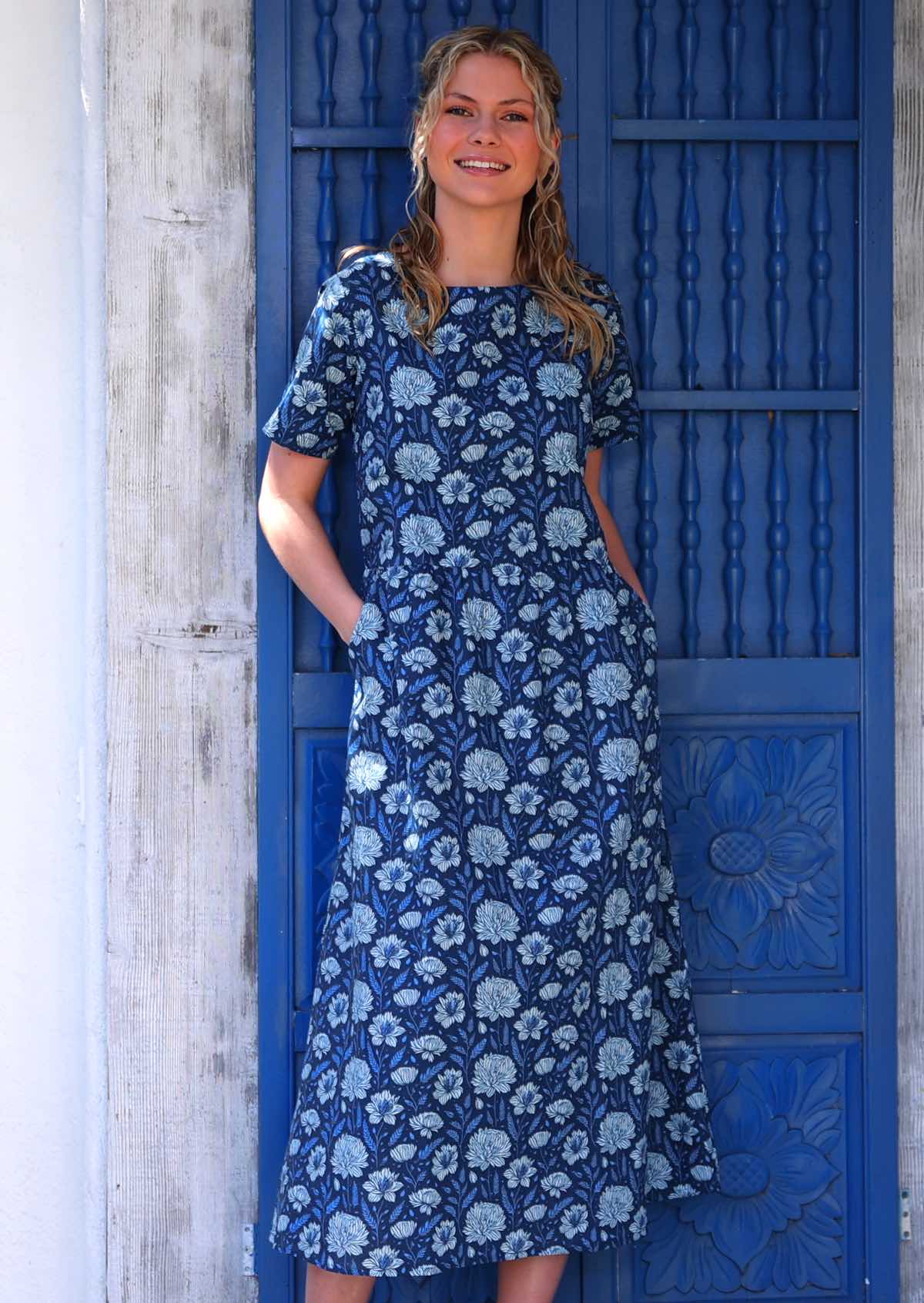 Model smiling standing in front of blue door with hands in pockets wearing pale floral on a navy base 100 percent cotton sundress