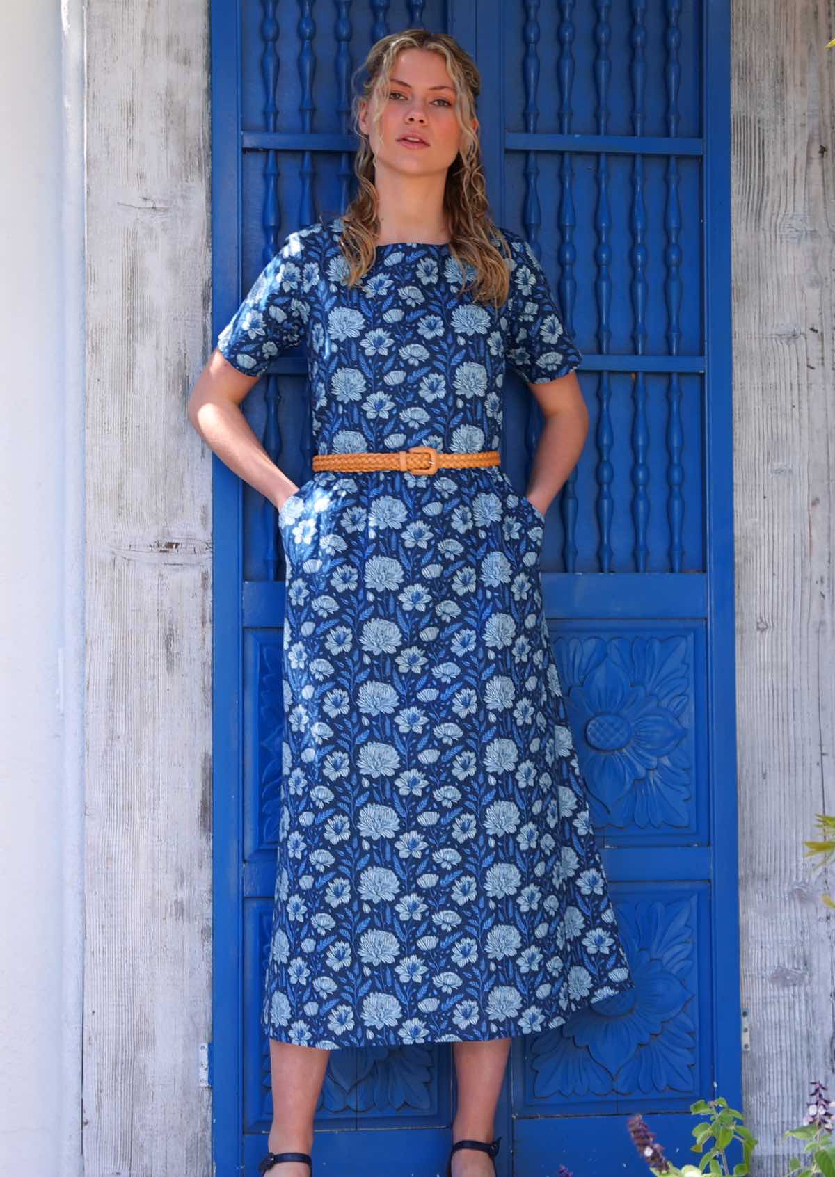 Model standing in front of blue door with hands in pockets wearing pale flowers on a navy base 100 percent cotton sundress paired with a leather waist belt 