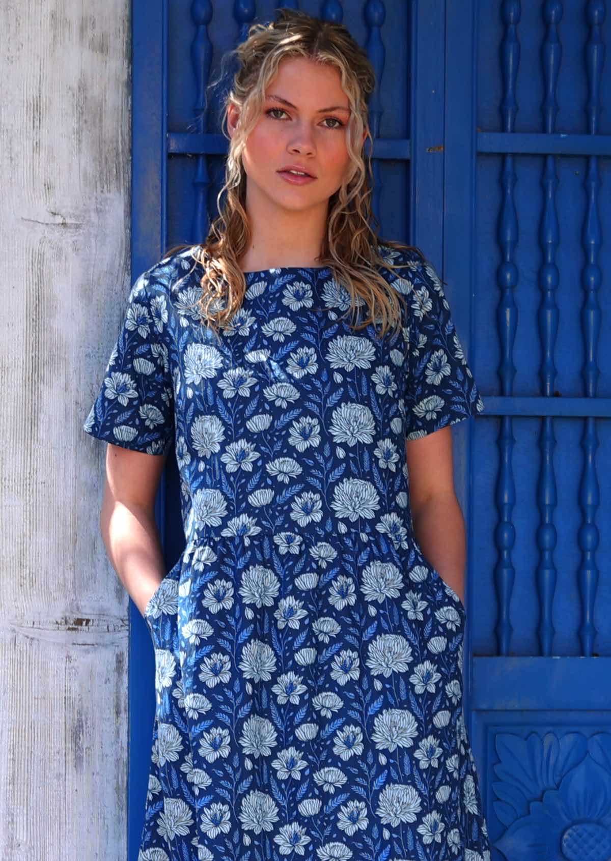 Model standing in front of blue door with hands in pockets wearing pale flowers on a navy blue base 100 percent cotton sundress