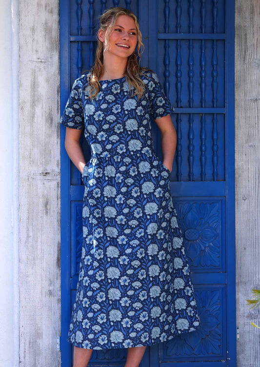 Model standing in front of blue door with hands in pockets wearing pale floral on a navy base 100 percent cotton sundress