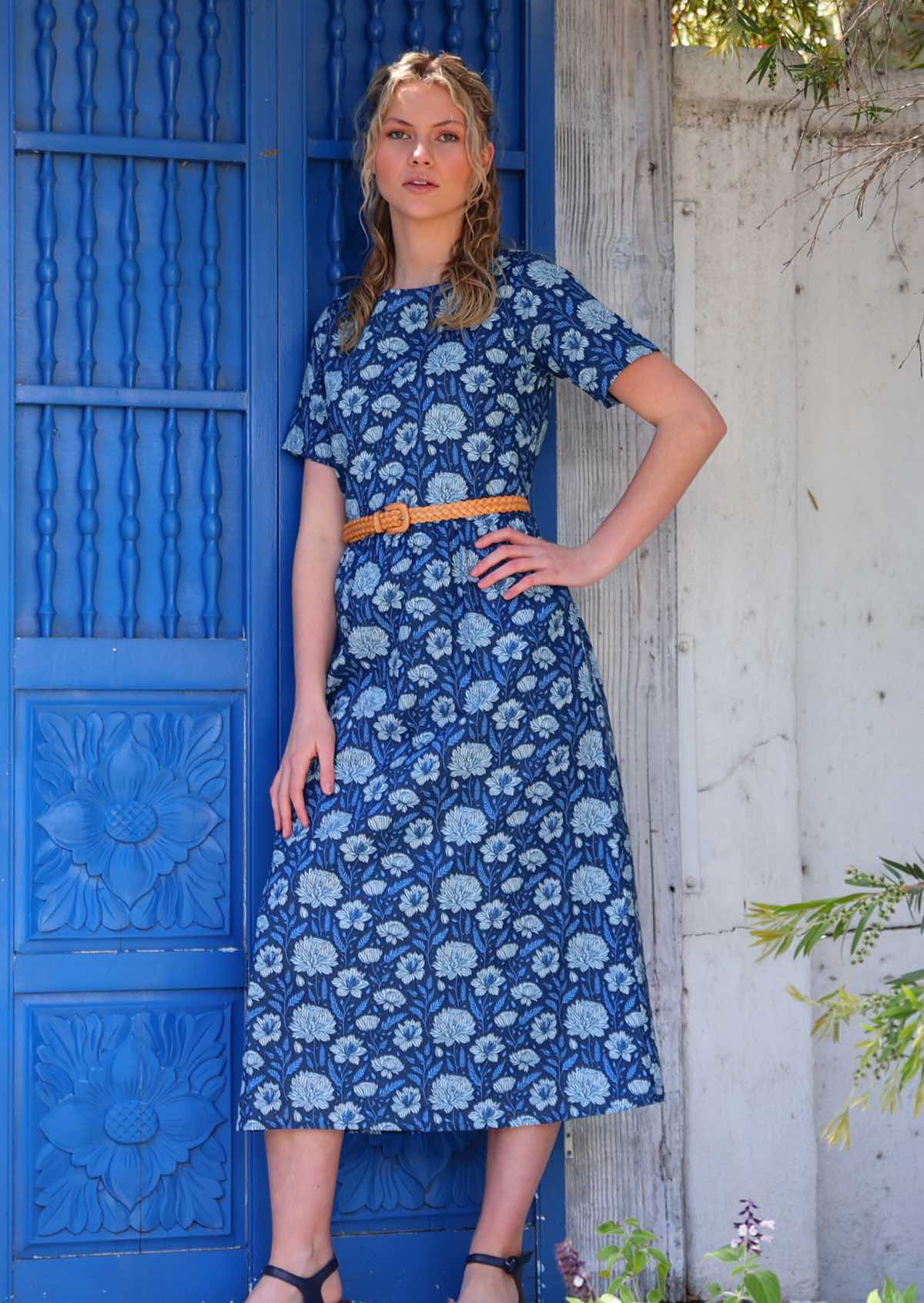 Model standing in front of blue door with hand on hip wearing pale flowers on a navy base 100 percent cotton sundress paired with leather waist belt