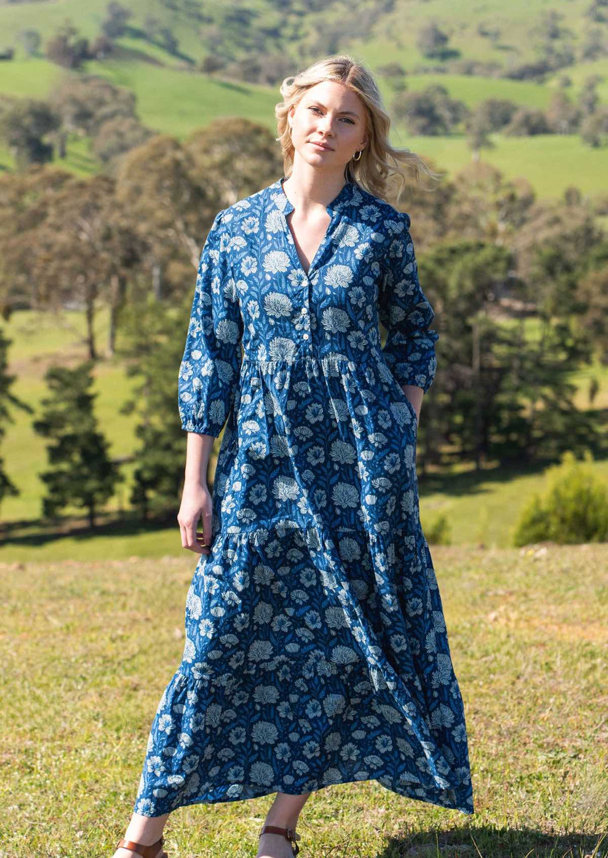 Model stands in field in cotton maxi dress with 3/4 sleeve and buttoned bodice, in blue based floral print