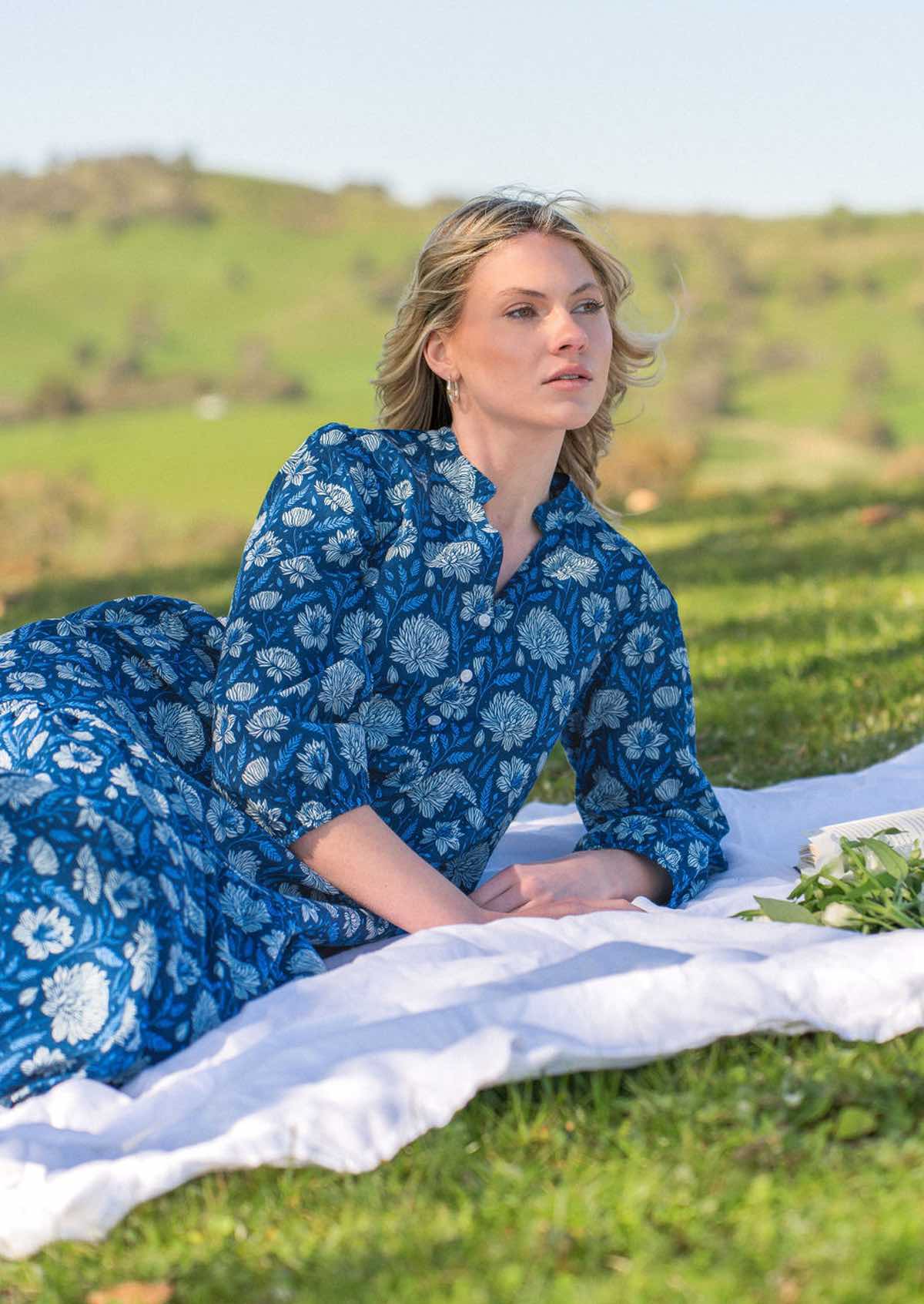 Model relaxes on picnic rug in blue floral cotton loose fit maxi dress with 3/4 sleeves and buttoned bodice