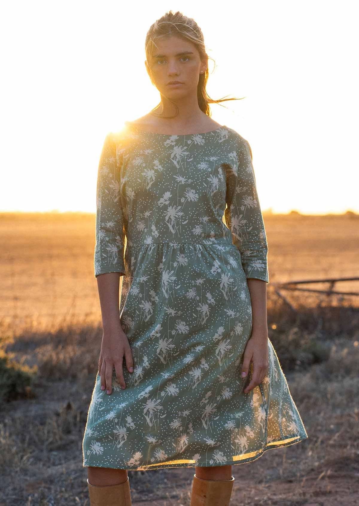 Model stands in setting sun light in a pale green based floral print cotton dress with 3/4 sleeves and boat neckline