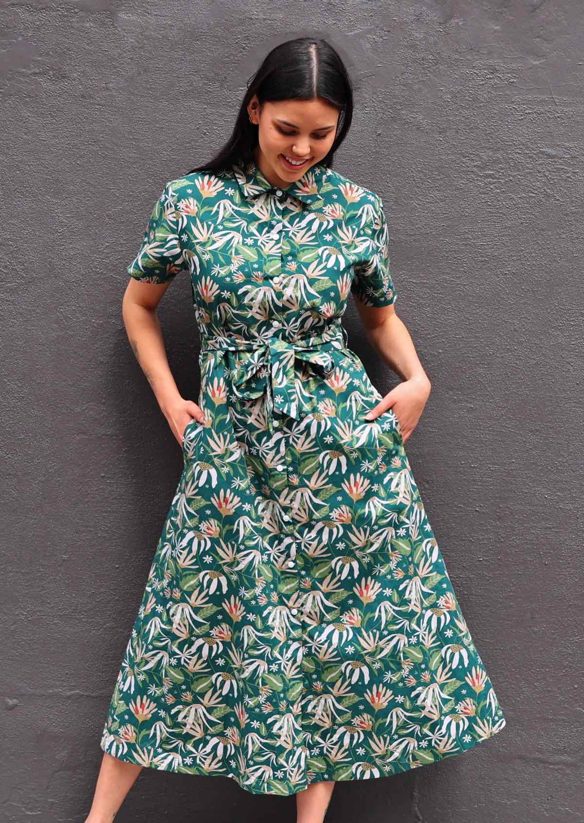 Woman with dark hair wearing 100% cotton green floral shirt dress, buttons up front, collar and pockets
