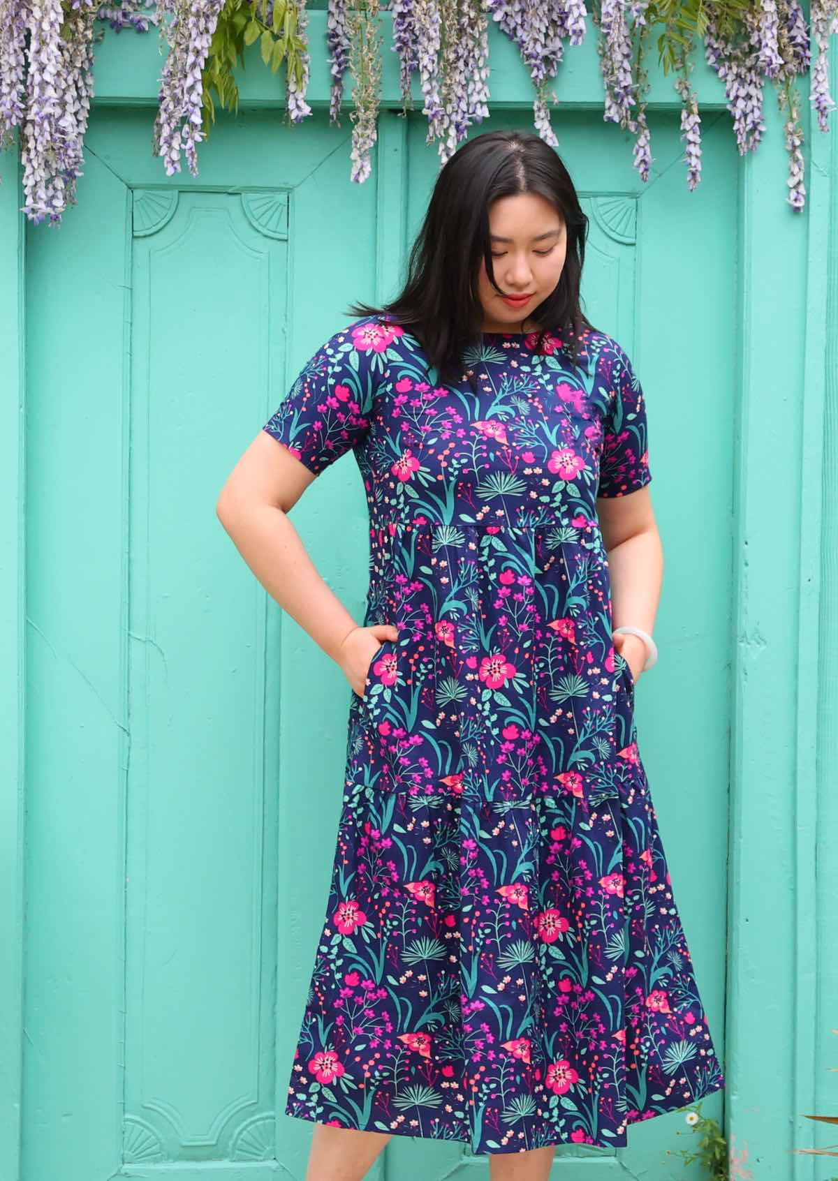 Model standing with hands in pockets wearing tiered long pink and purple floral on dark blue base 100 percent cotton sundress 