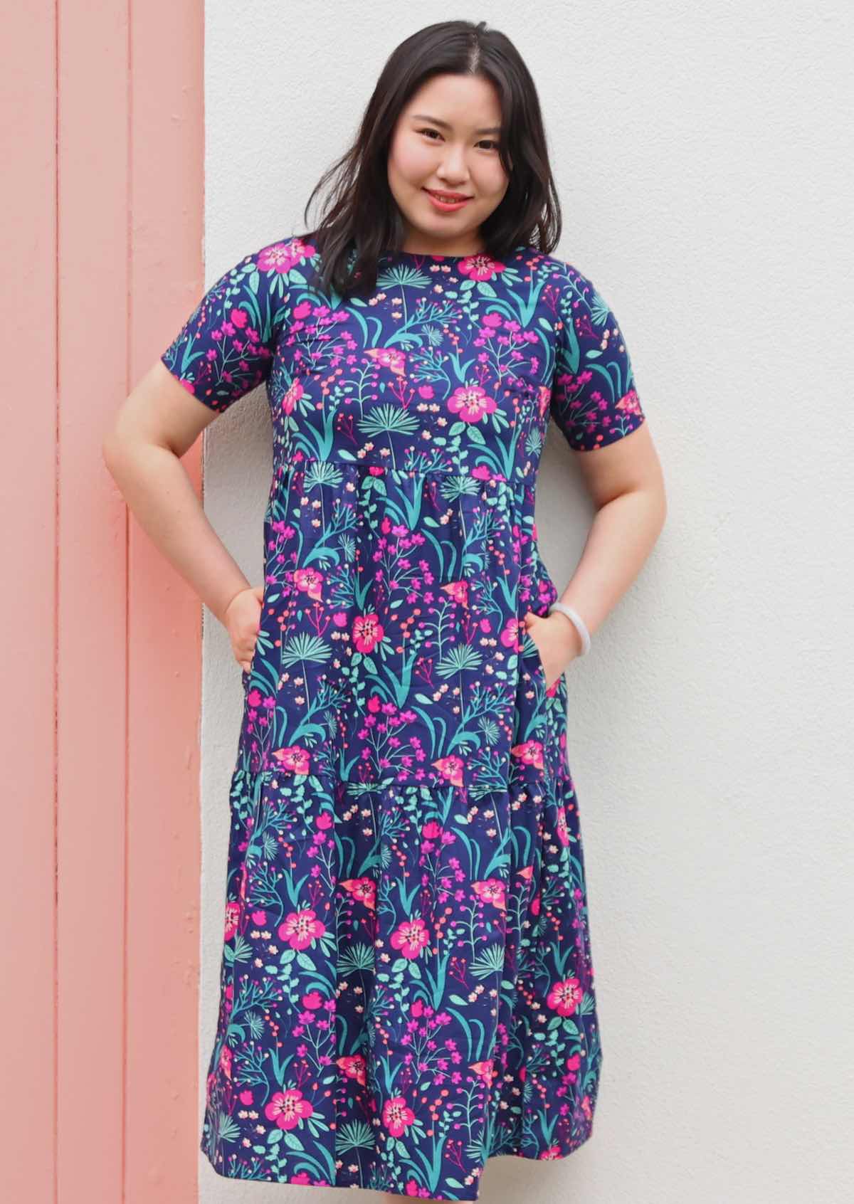 Model standing and smiling with hands in pockets wearing tiered long pink and purple floral on dark blue base 100 percent cotton sundress 