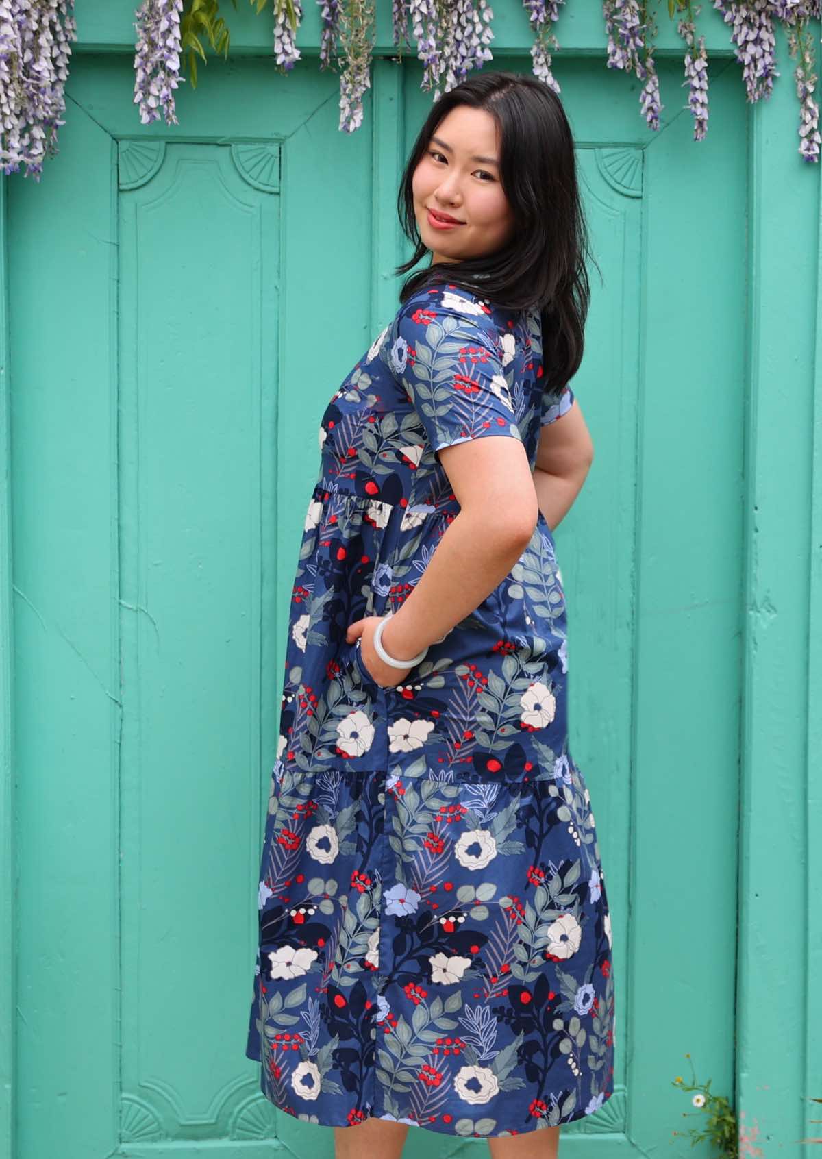 Woman with dark hair wearing a blue 100% cotton dress with white and blue floral design, in front of aqua door