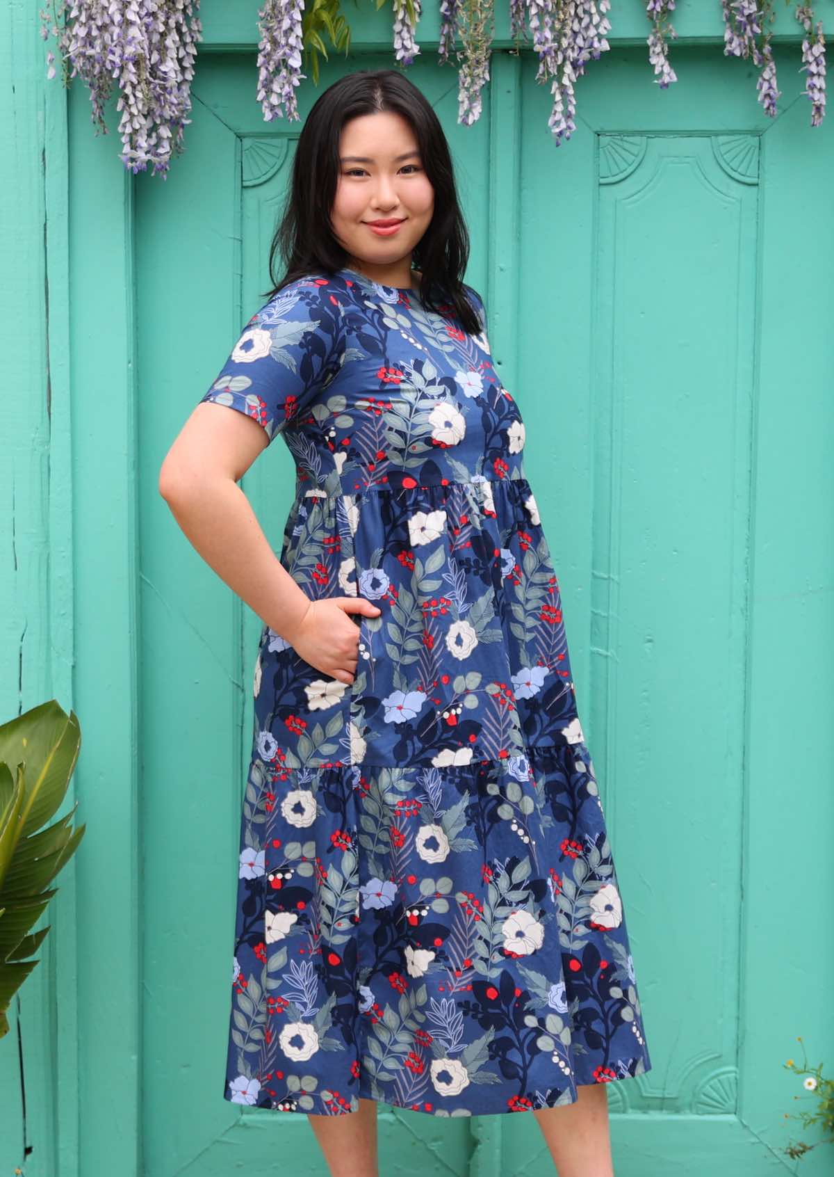 Woman with dark hair wearing a  blue floral cotton dress with white and blue flowers, in front of aqua door