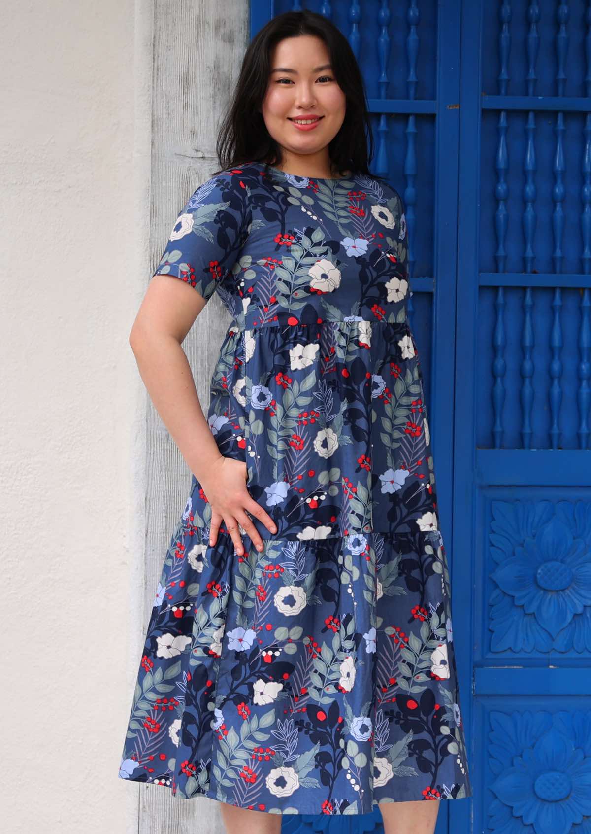 Woman with dark hair wearing a  blue floral cotton dress with white and blue flowers, with pockets in front of blue door