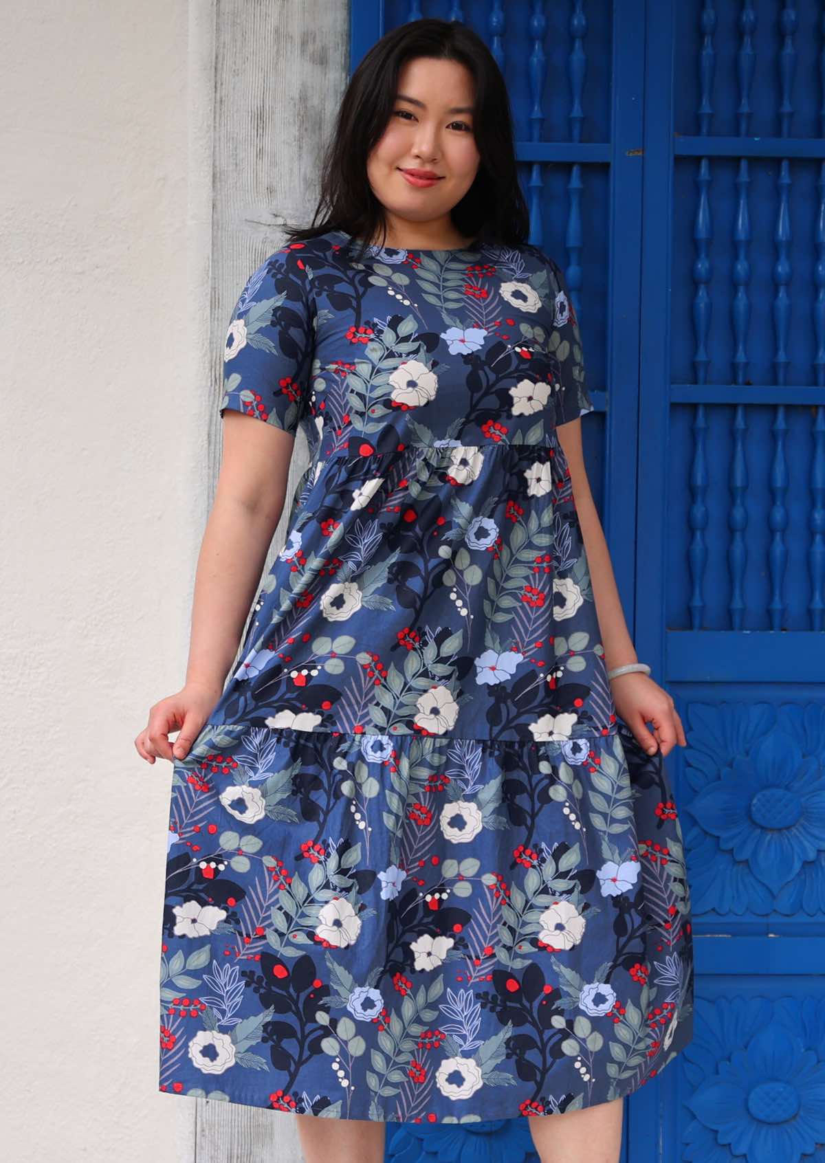 Woman with dark hair wearing a blue cotton dress with white and blue floral design, in front of blue door