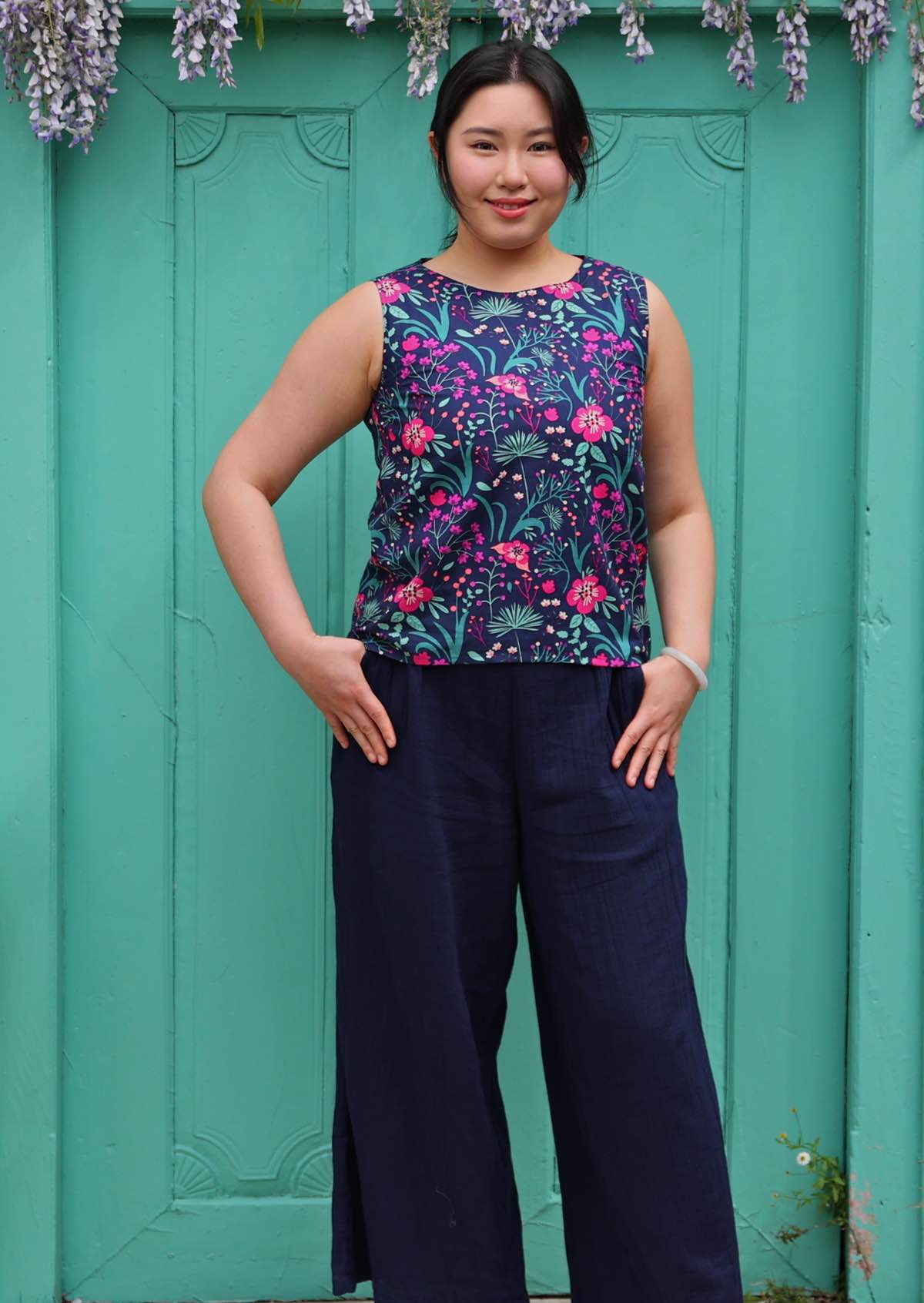 Model standing in front of aqua door with thumbs in pockets 100 percent cotton pink and purple florals on a dark blue base sleeveless top
