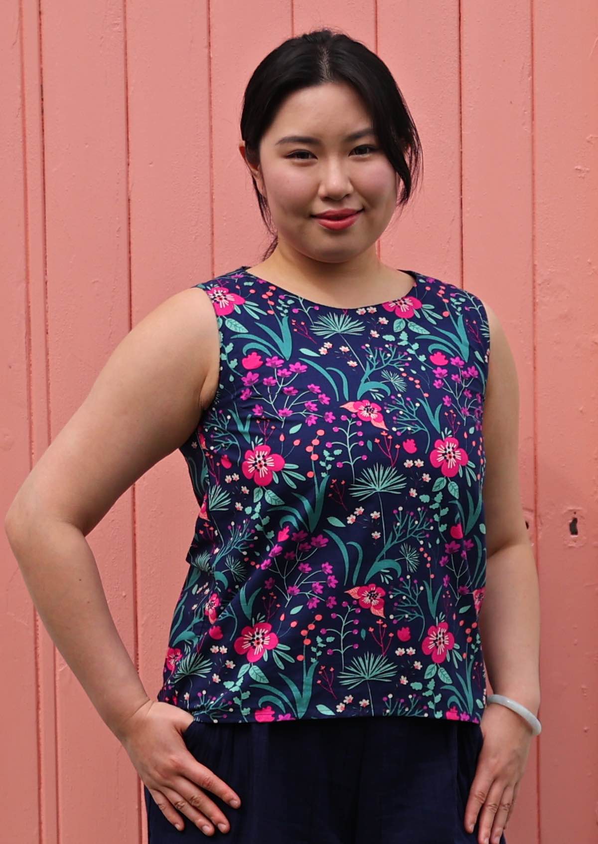 Model standing in front of pink door with thumbs in pockets 100 percent cotton pink and purple florals on a dark blue base sleeveless top