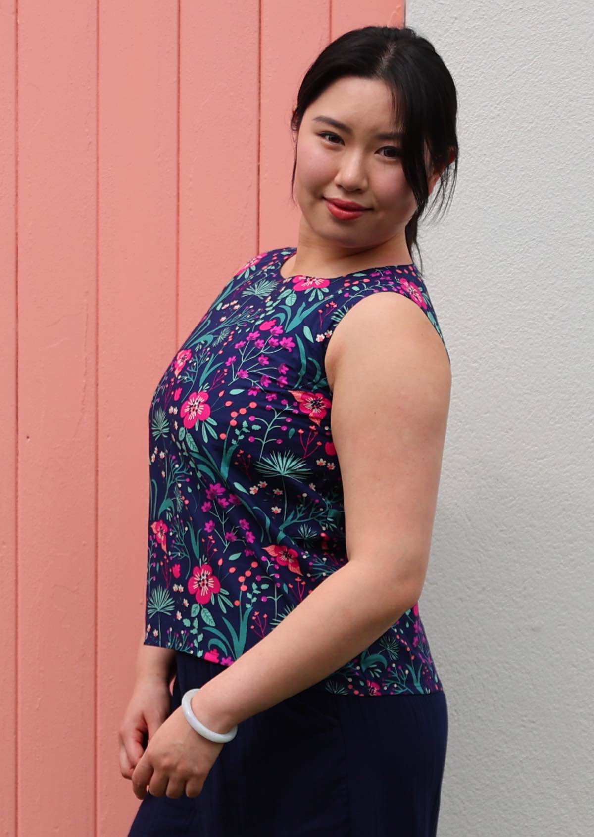 Model standing in front of pink door wearing 100 percent cotton pink and purple florals on a dark blue base sleeveless top