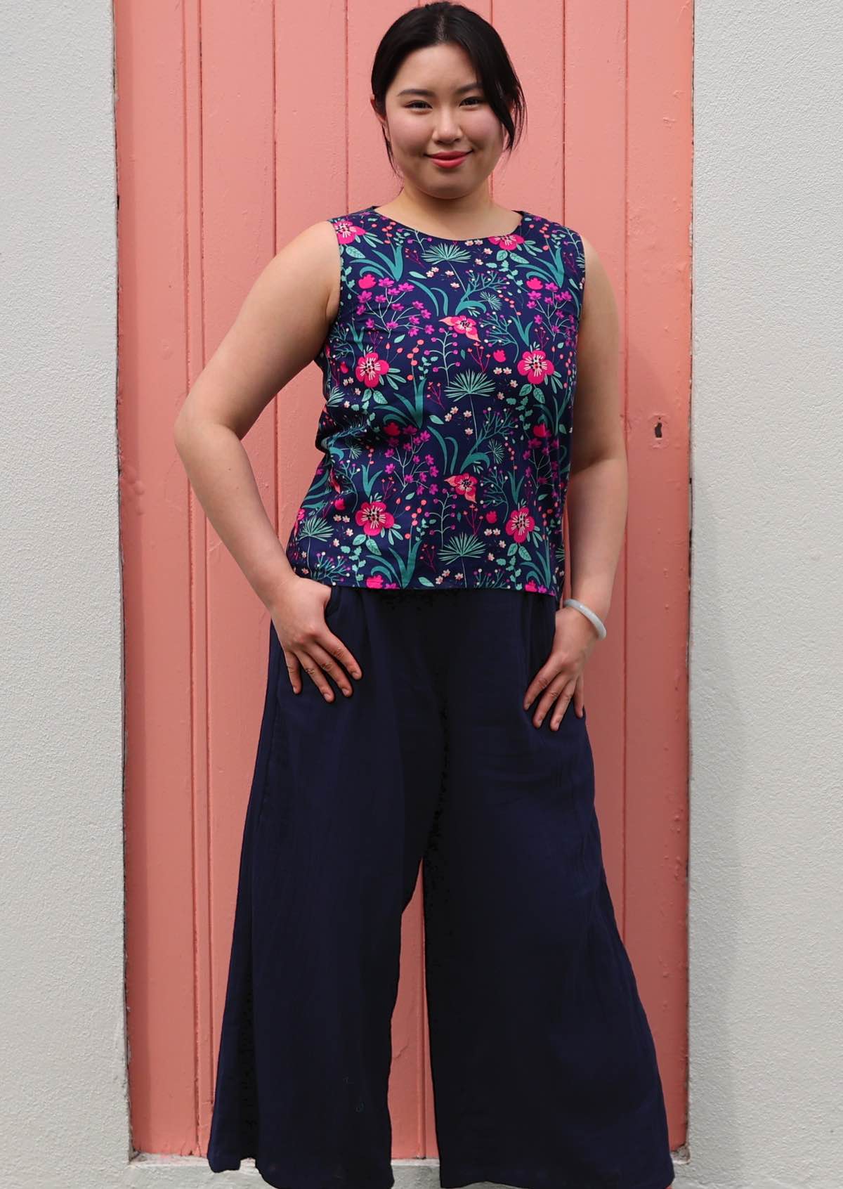 Model smiling standing in front of pink door with thumbs in pockets 100 percent cotton pink and purple florals on a dark blue base sleeveless top