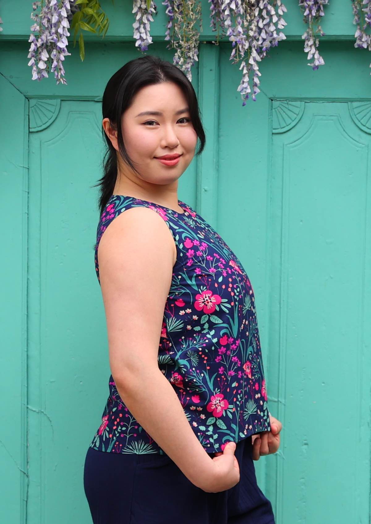 Model standing in front of aqua door wearing 100 percent cotton pink and purple florals on a dark blue base sleeveless top