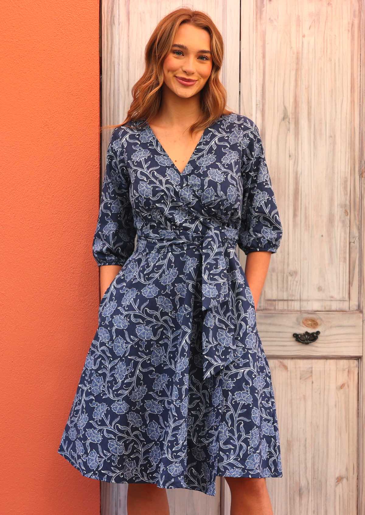 Model standing in front of door wearing Ella Wrap Dress Neela 100 percent cotton 3/4 sleeve navy blue floral wrap dress with hands in pockets