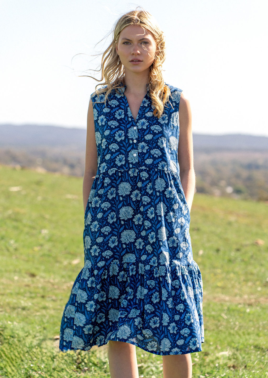 Woman stands in field in cotton sleeveless midi length dress in blue floral print with hands in deep hidden side pockets