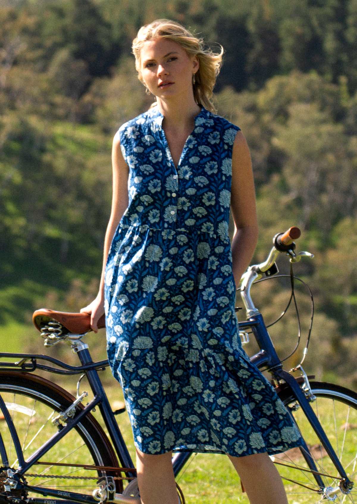 Woman stands in front of bicycle in cotton sleeveless midi length dress with V-neck and mandarin collar, in blue floral print