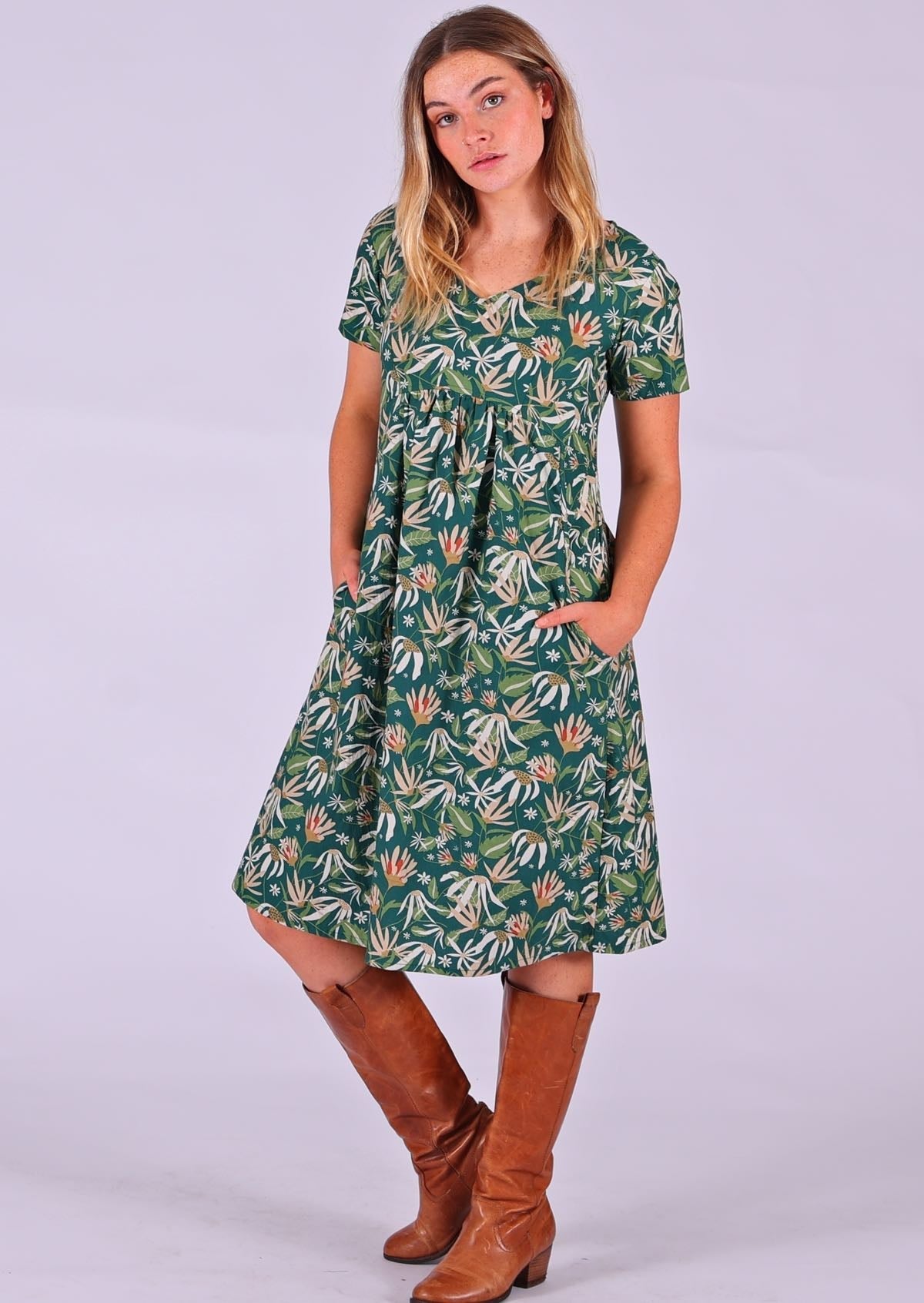 Model standing with hands in pockets modelling the Etta v-neck Dress Flannel Flower 100 percent cotton green sundress with short sleeves paired with tan leather boots