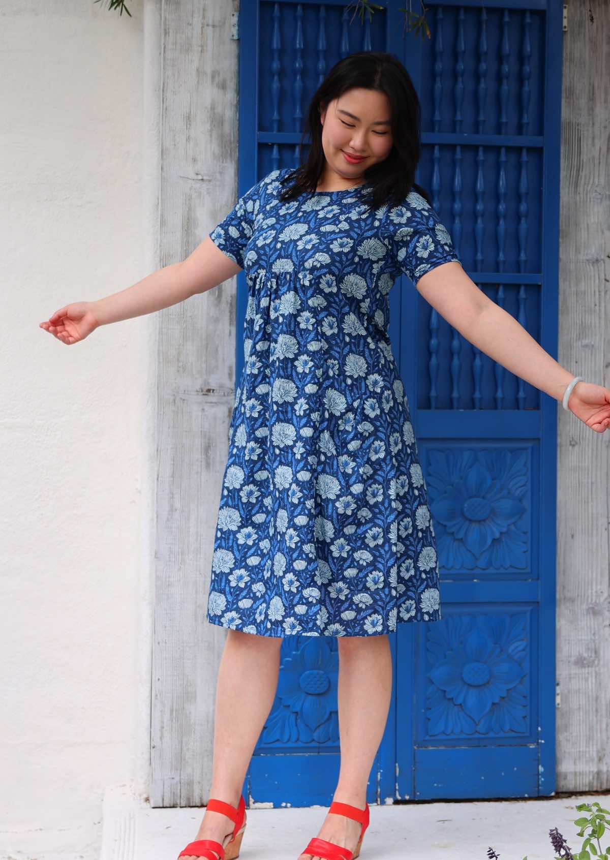 Woman with dark hair wearing a blue cotton knee length dress with light blue and white floral design, in front of blue door