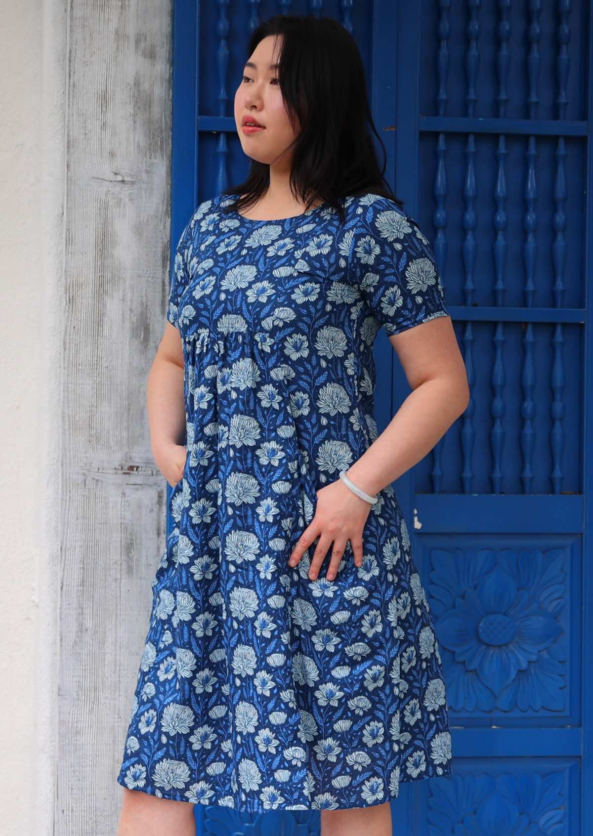 Model in blue floral cotton knee length dress with short sleeves and empire waistline, with hands in pockets