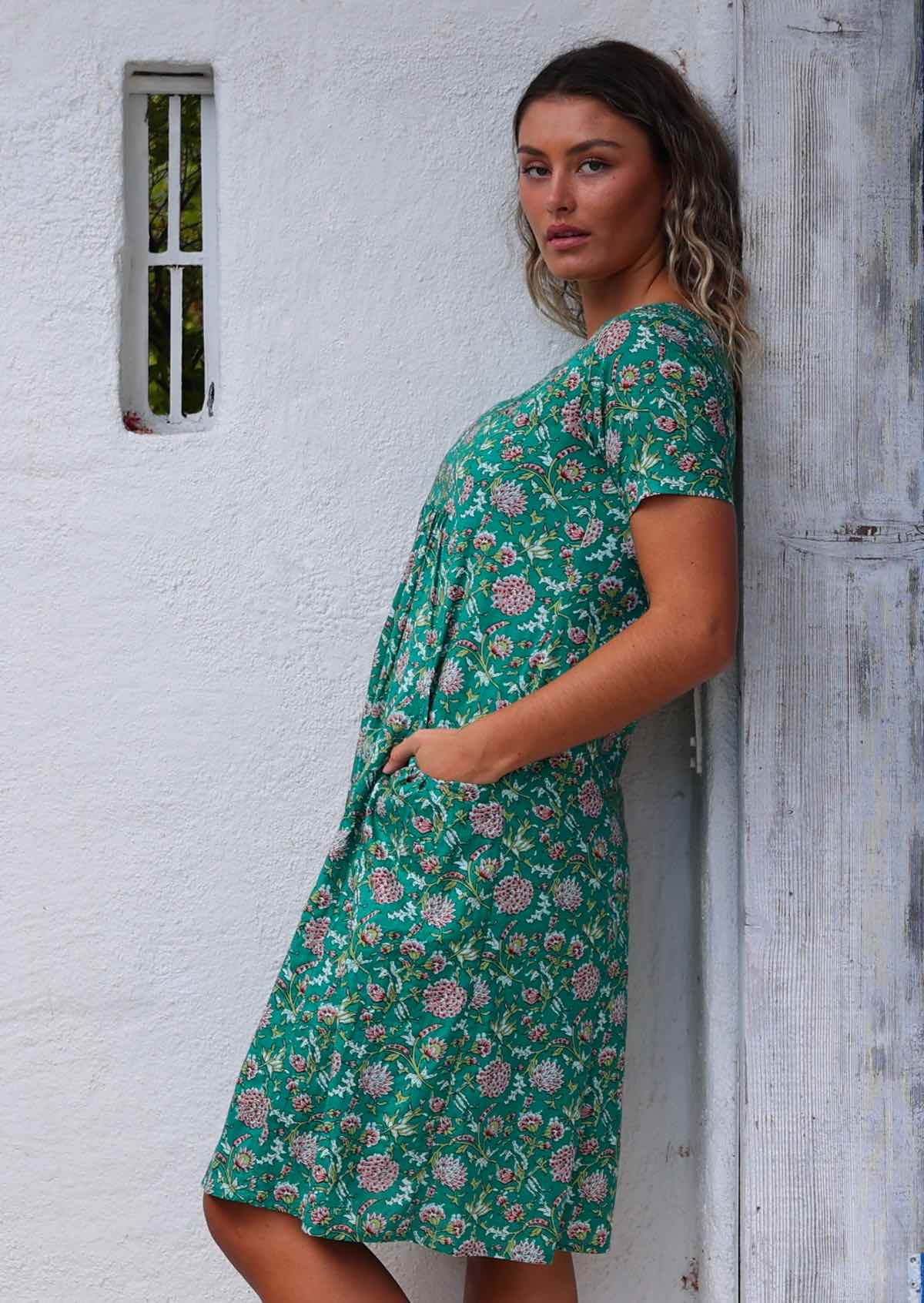 Model leaning on wooden post with hand in pocket wearing 100 percent cotton green sundress 