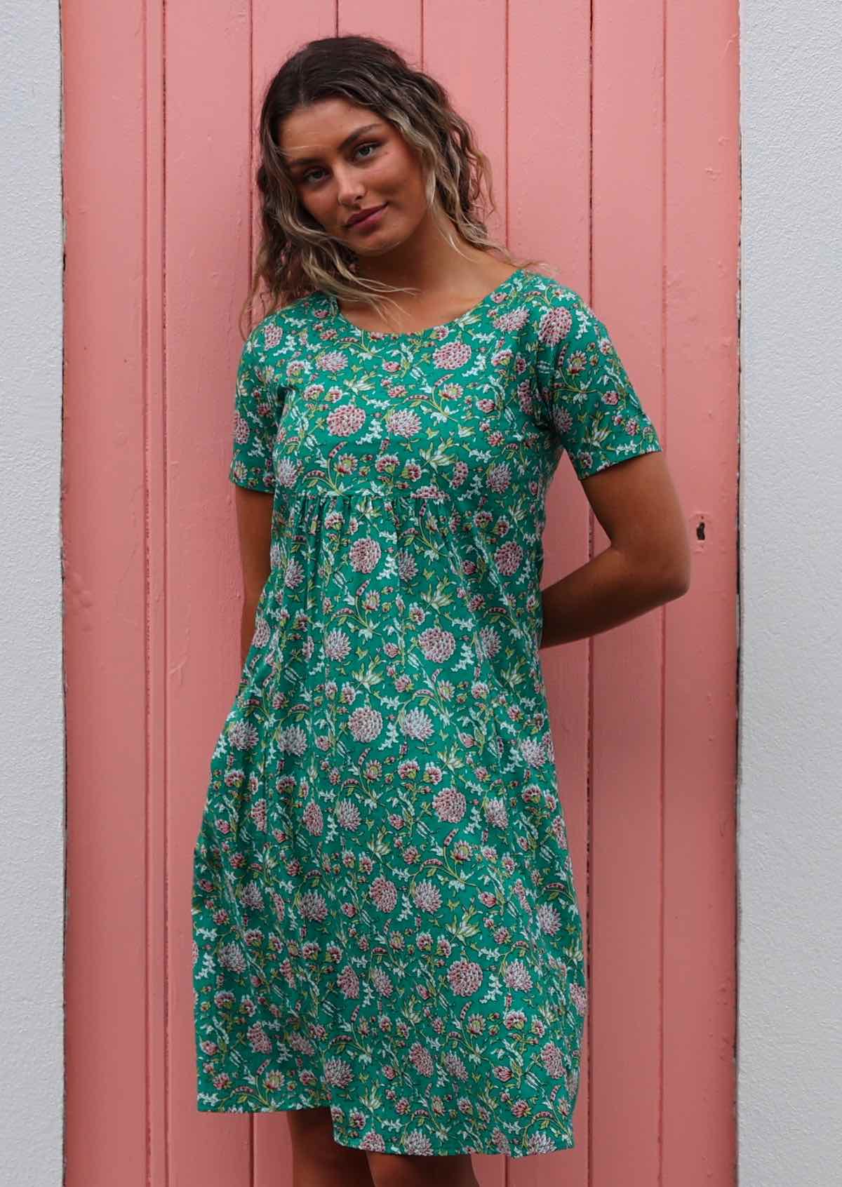 Model standing in front of pink door with hands behind back wearing 100 percent cotton green sundress 