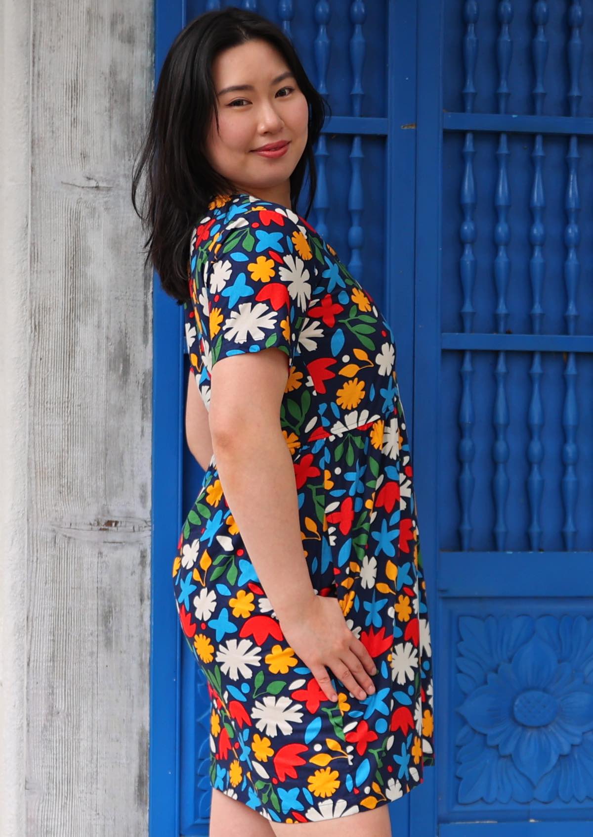 Woman stands turned to the side in above knee cotton dress in bold floral print with thumb in pocket