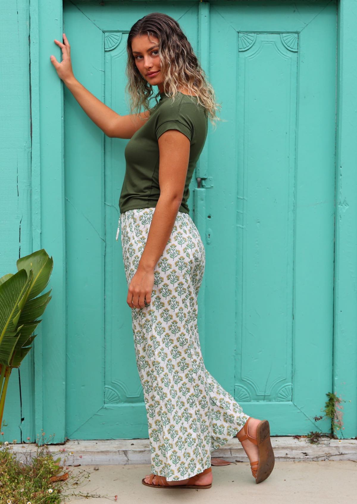 side view of woman wearing white cotton palazzo pant with floral detail in front of aqua door 