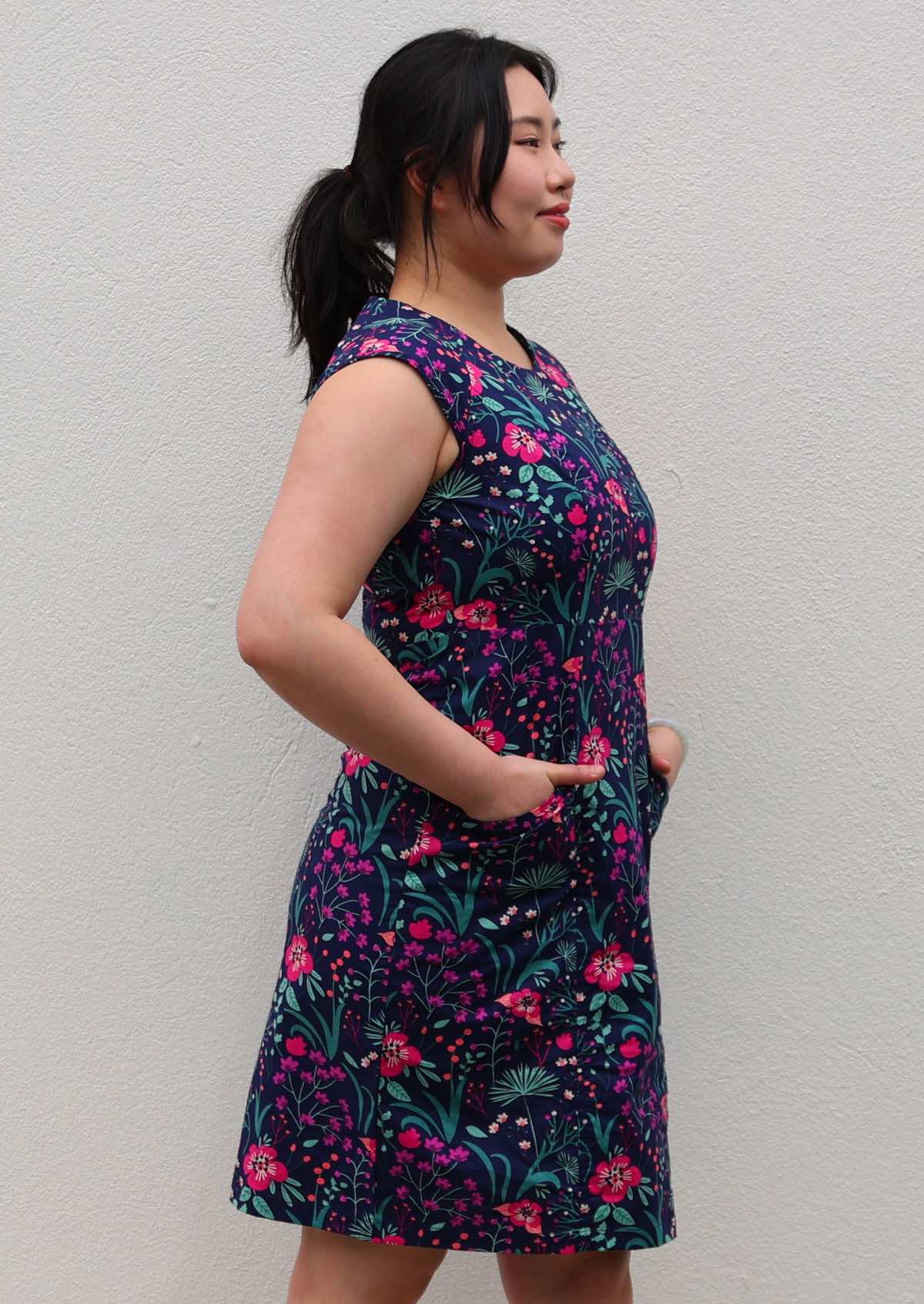 Woman with dark hair in 100% navy cotton floral dress with hands in pockets side view
