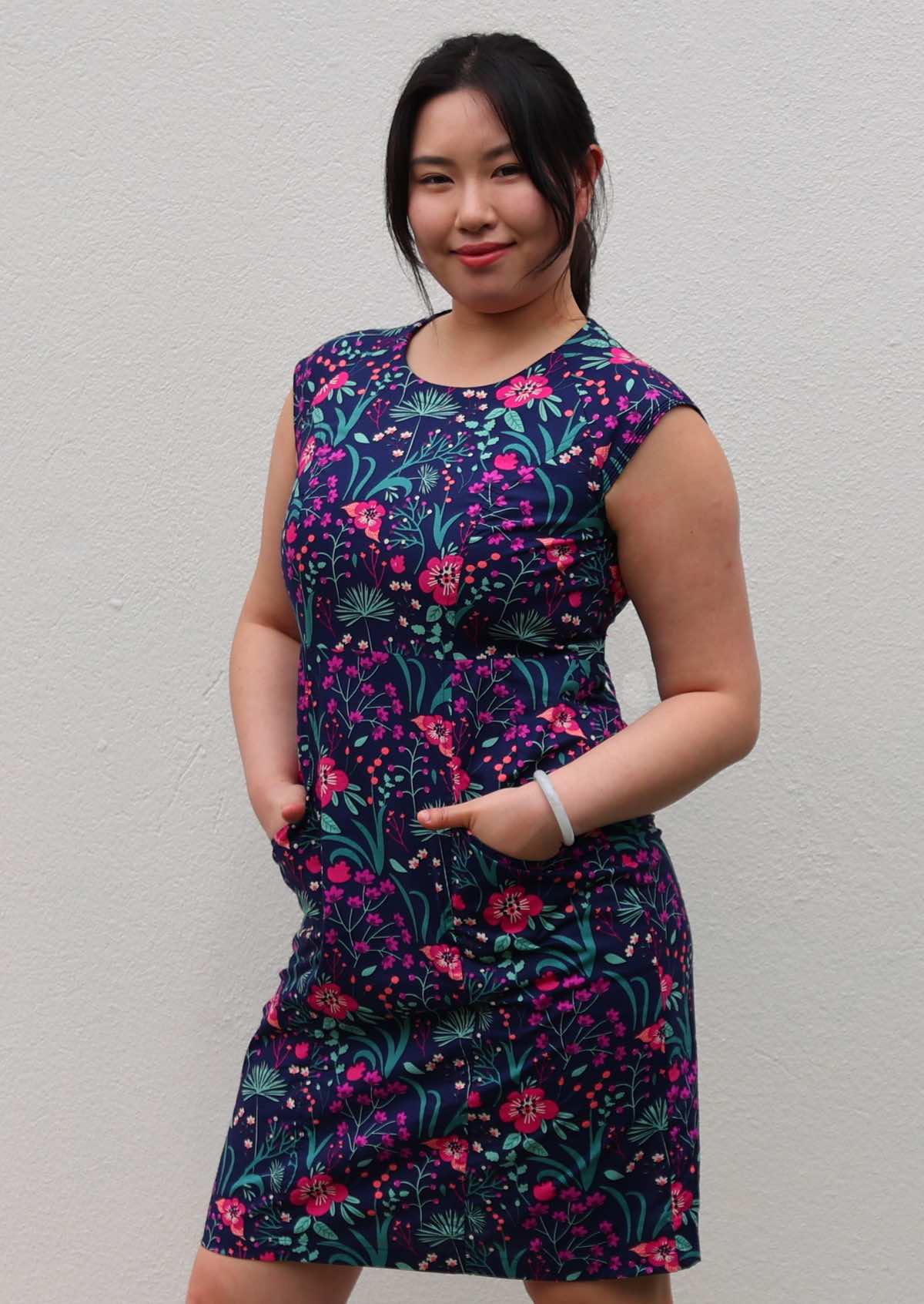 Woman with dark hair in 100% navy cotton floral dress with hands in pockets in front of white wall
