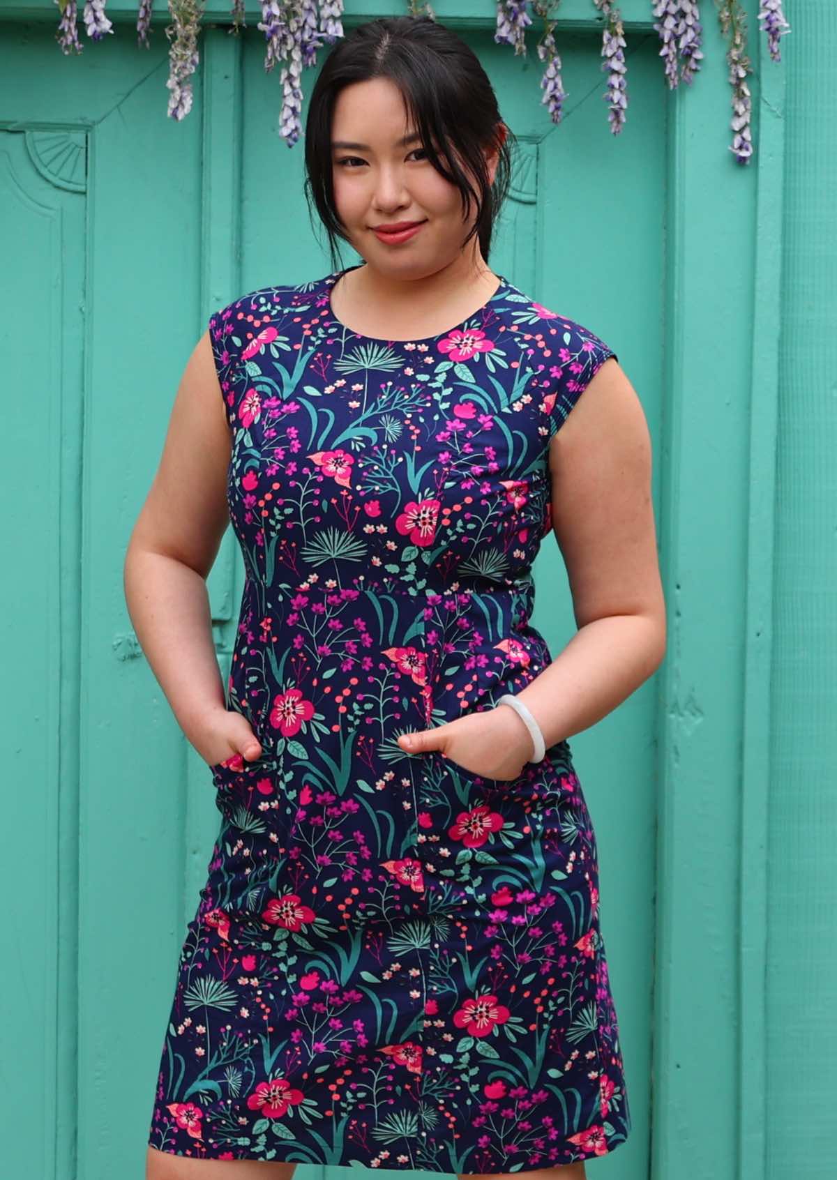 Woman with dark hair in 100% navy cotton floral dress with hands in pockets underneath wisteria