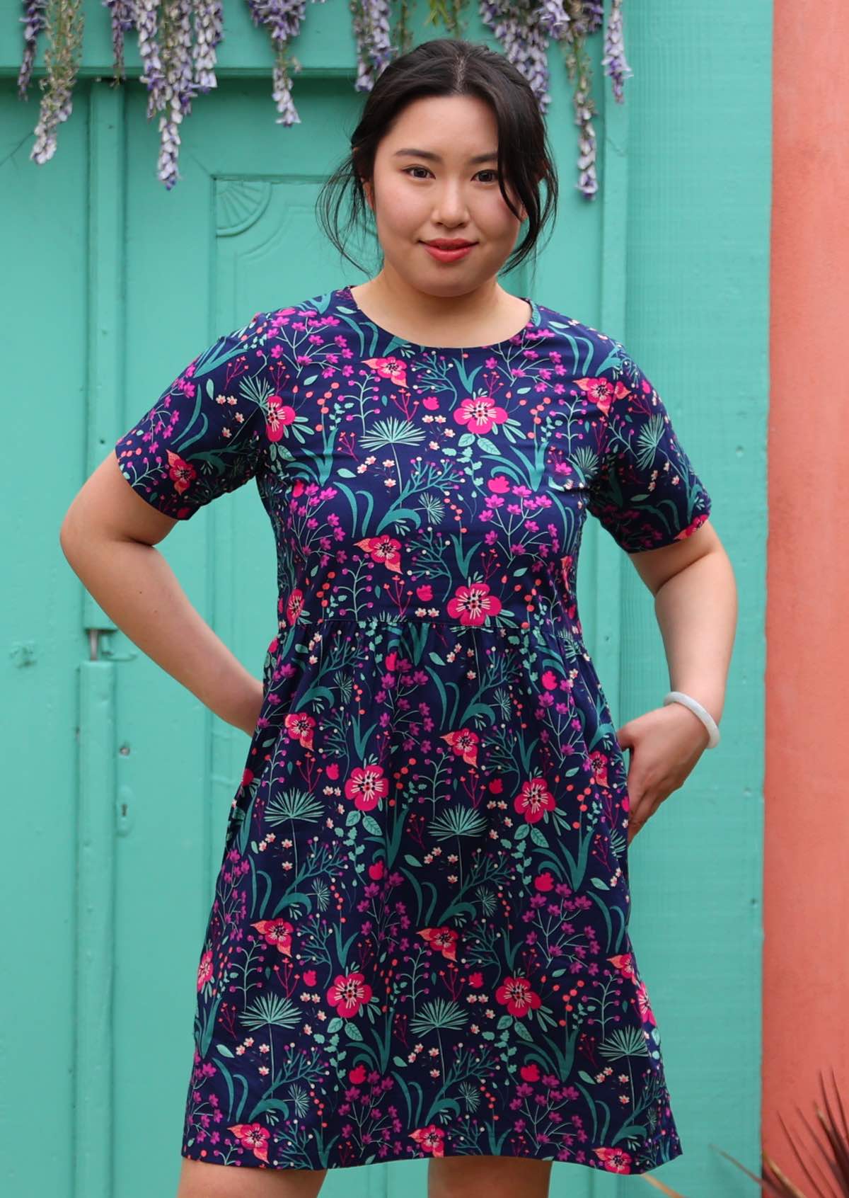 woman in loose fit purple, navy and pink floral print 100% cotton dress in front of aqua door hands in pockets