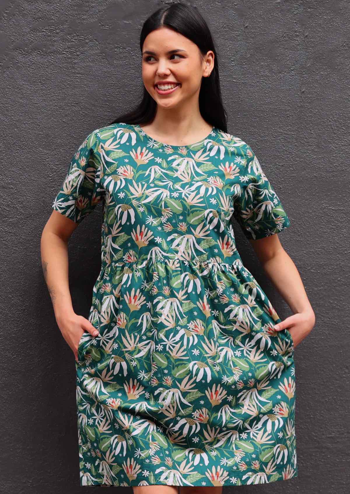 Model with dark hair in loose fit green cotton floral dress with hands in pockets in front of dark grey wall