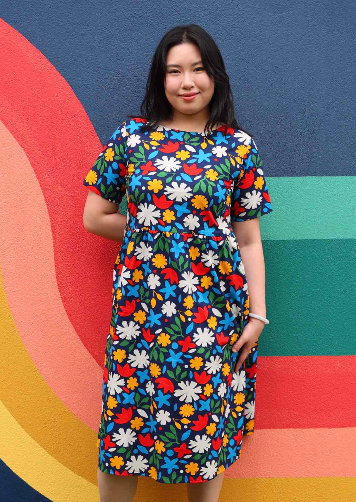 Model leans on wall in cotton midi length relaxed fit dress in bold floral print on a dark blue base