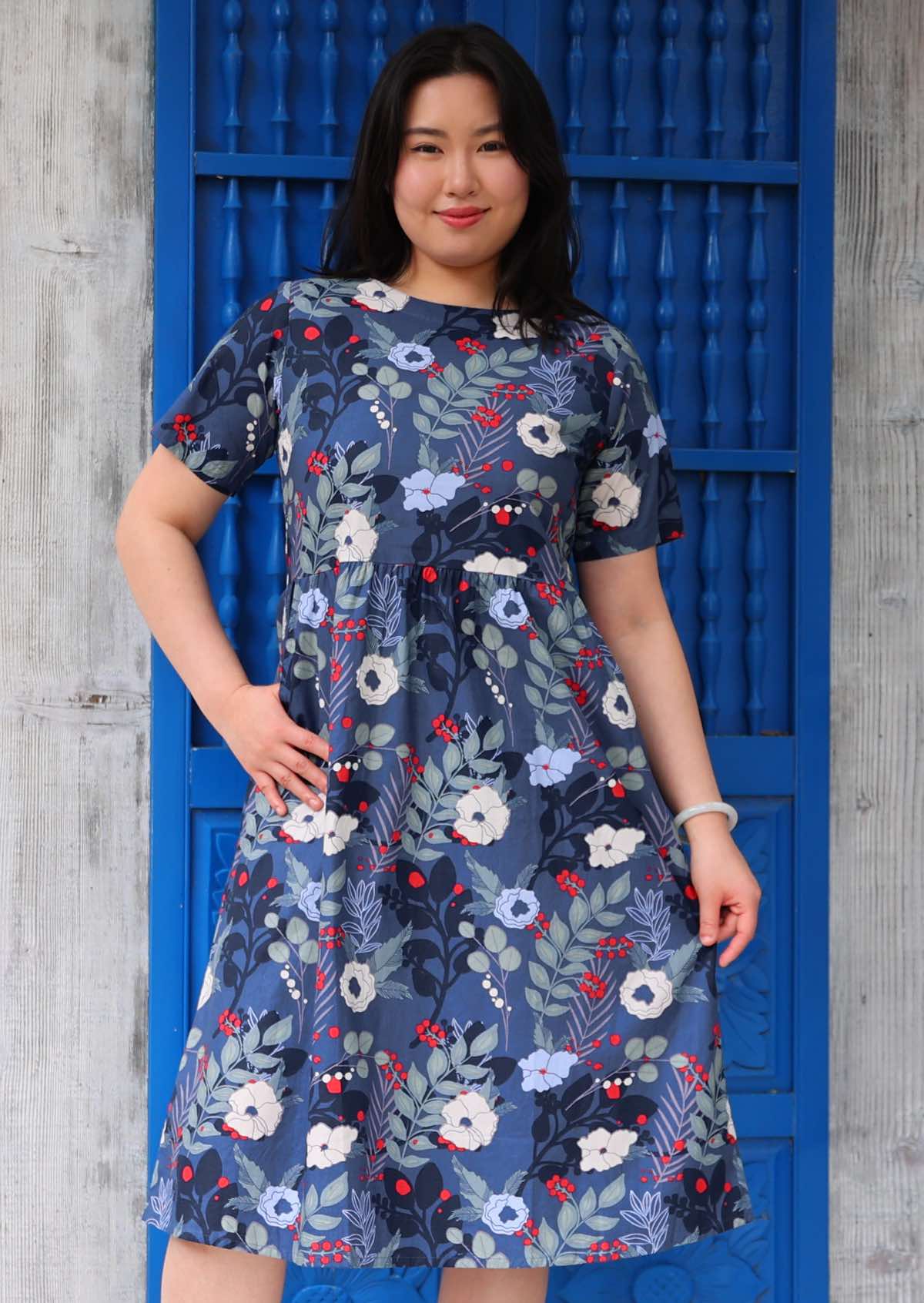 Woman with dark hair wearing a blue cotton short sleeved dress with white and blue floral print with pops of red, in front of blue door