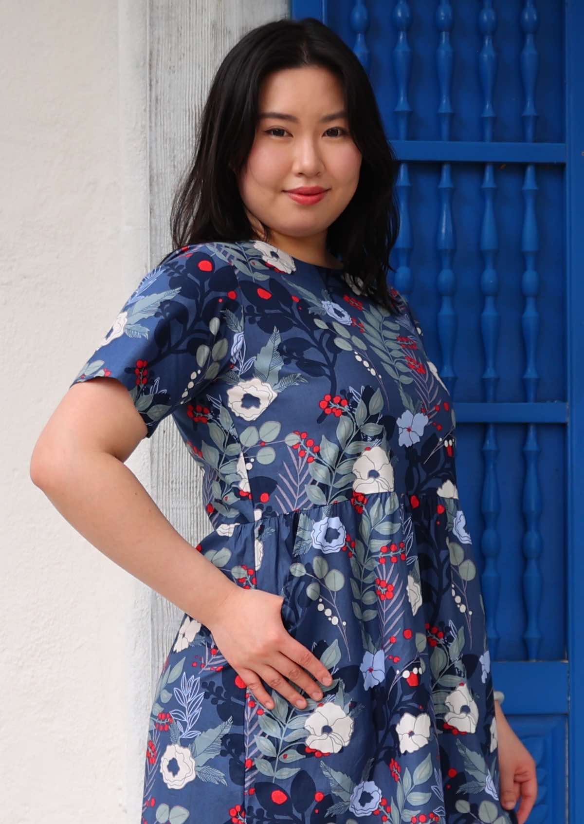 Woman with dark hair wearing a blue cotton dress with white and blue floral design, in front of blue door, with thumb in side pocket