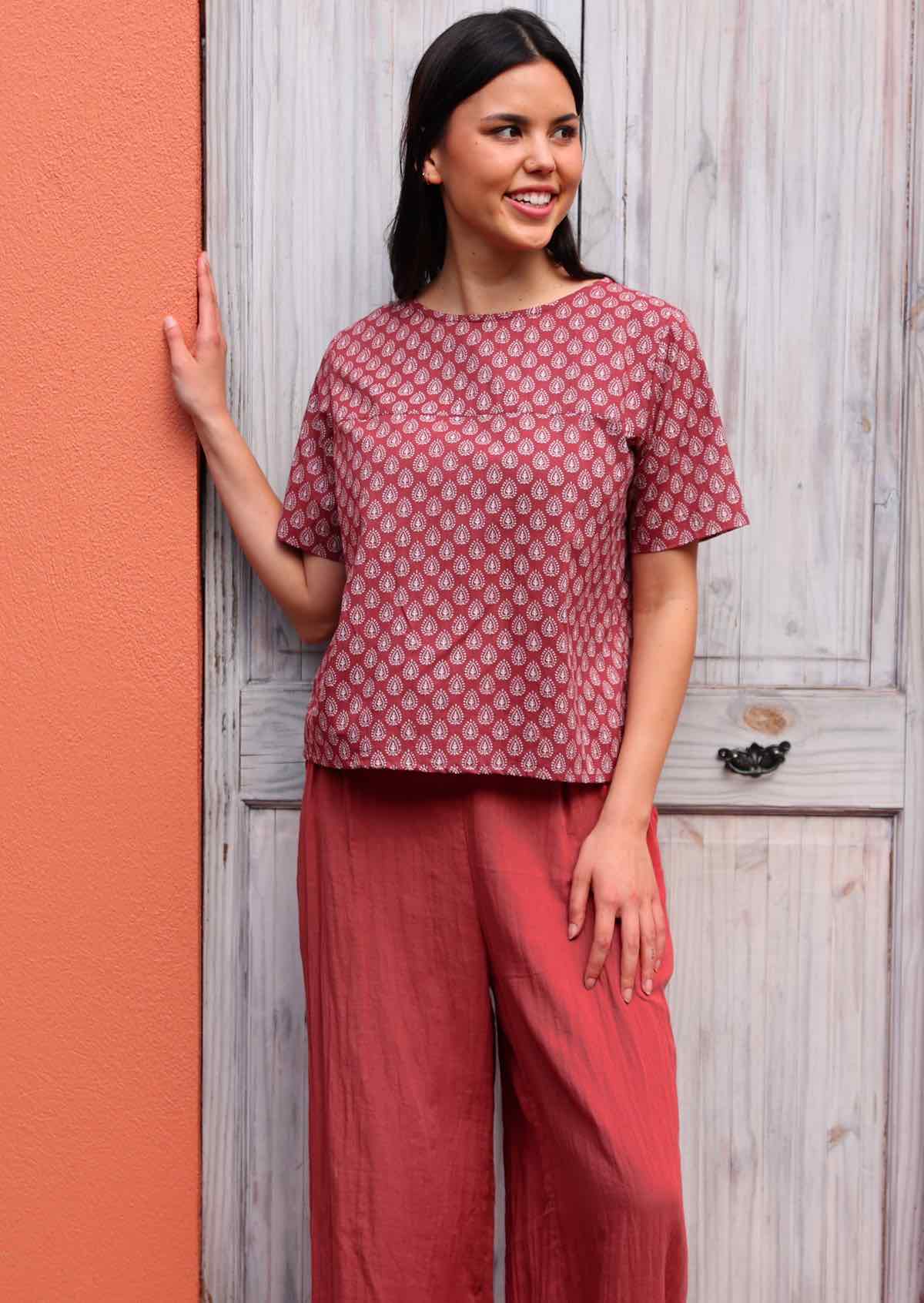 Model leans on door frame in boxy style cotton top in dark terracotta and white print, paired with terracotta cotton pants