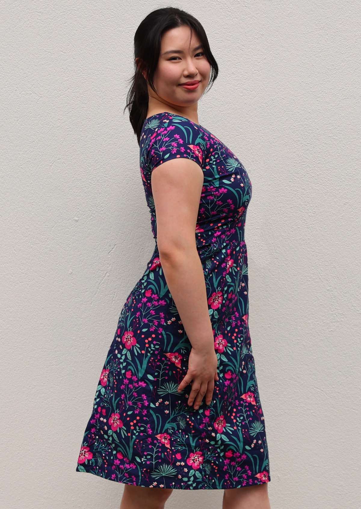 woman in navy and purple floral cotton knee length dress with round neckline and hands in pockets in front of white wall side view
