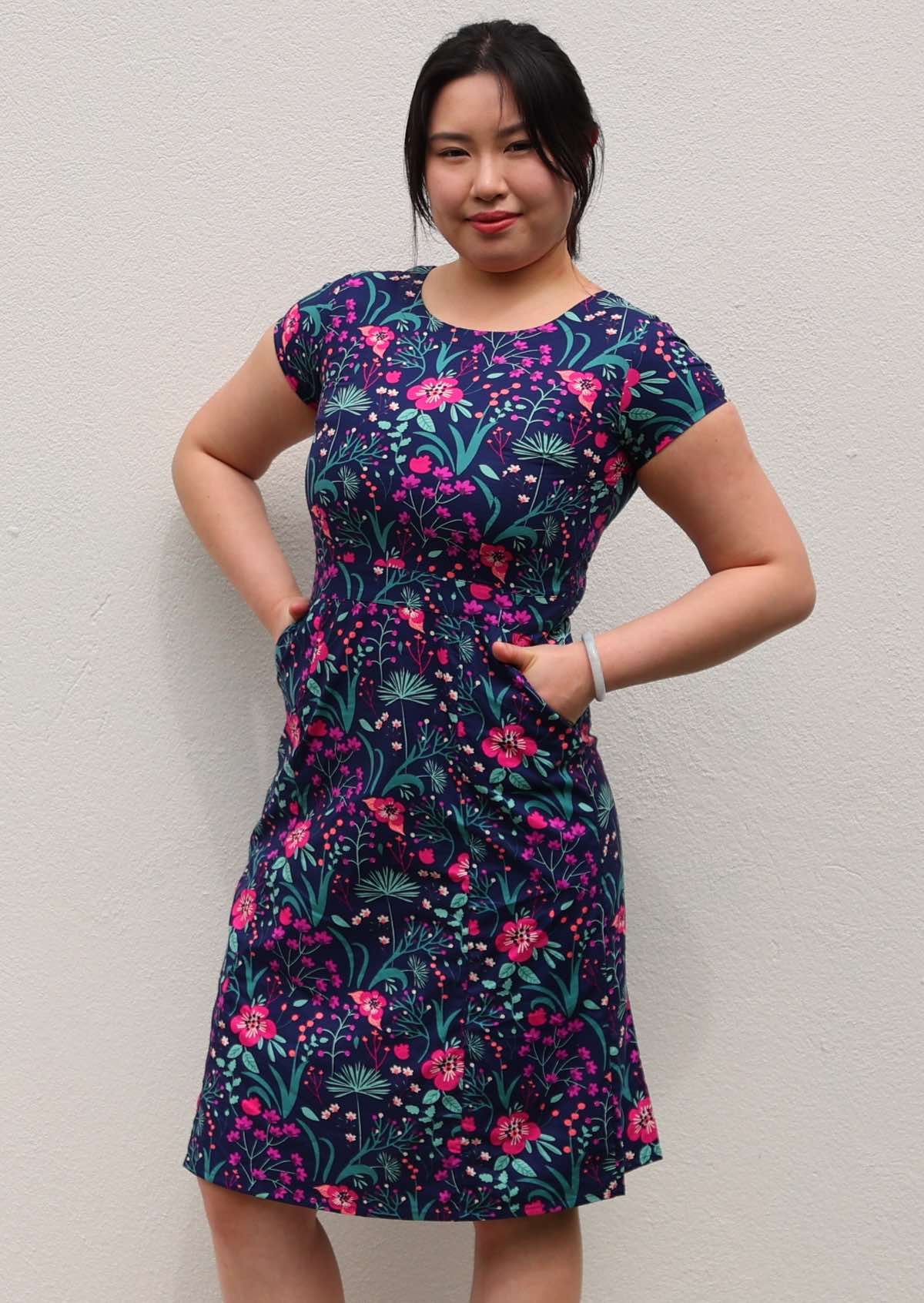 woman in navy and purple floral cotton knee length dress with round neckline and hands in pockets in front of white wall 