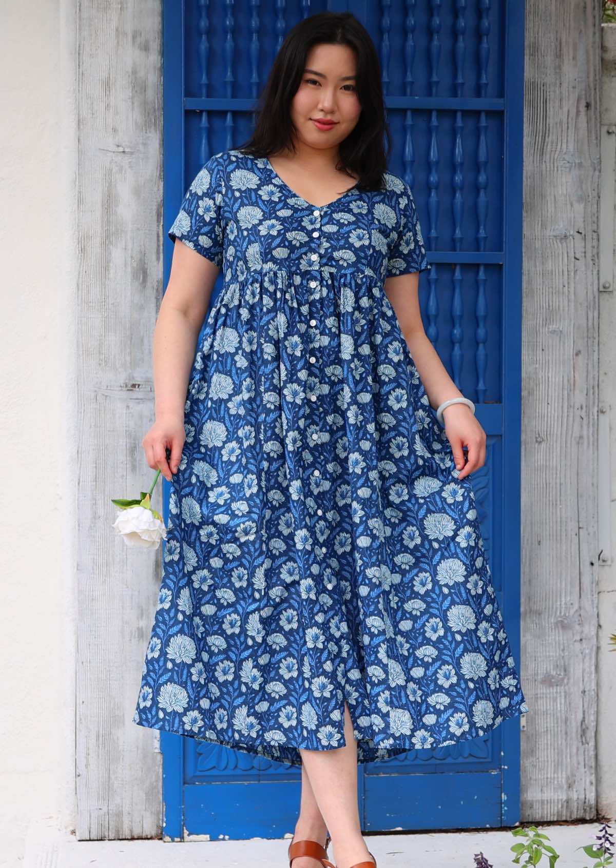 Model standing in front of blue door holding a flower wearing 100 percent cotton navy blue button through v-neck dress with pockets