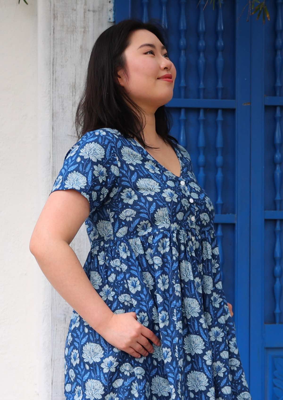 Model standing in front of blue door with thumbs in pockets wearing 100 percent cotton navy blue button through v-neck dress 