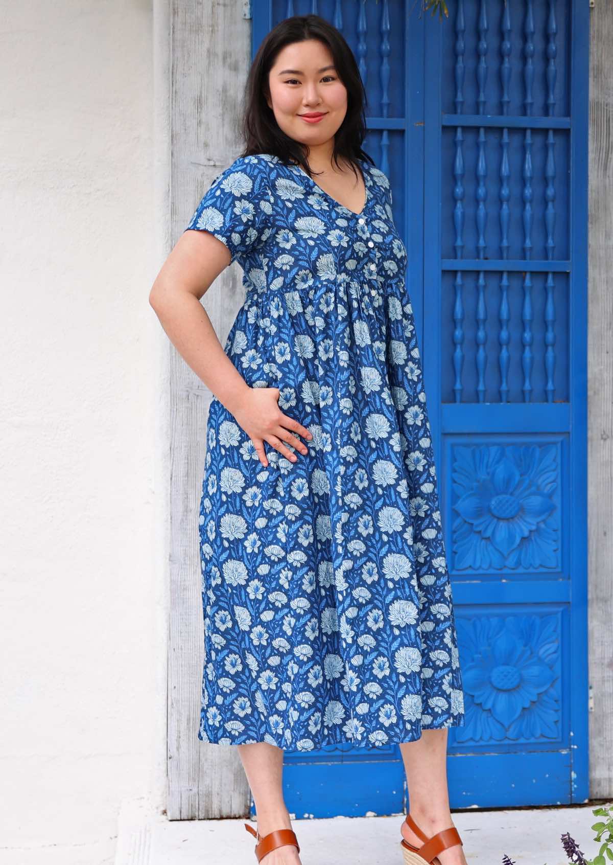 Model standing side on in front of blue door with thumb in pocket wearing 100 percent cotton navy blue button through v-neck dress with pockets