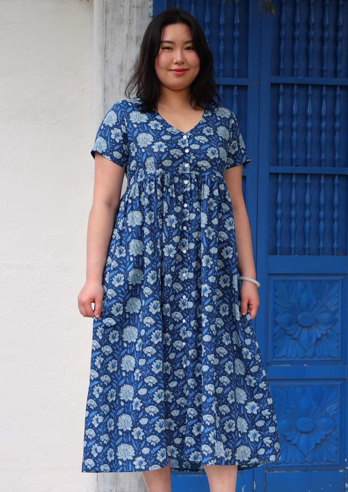 Model standing and smiling in front of blue door wearing 100 percent cotton navy blue button through v-neck dress with pockets