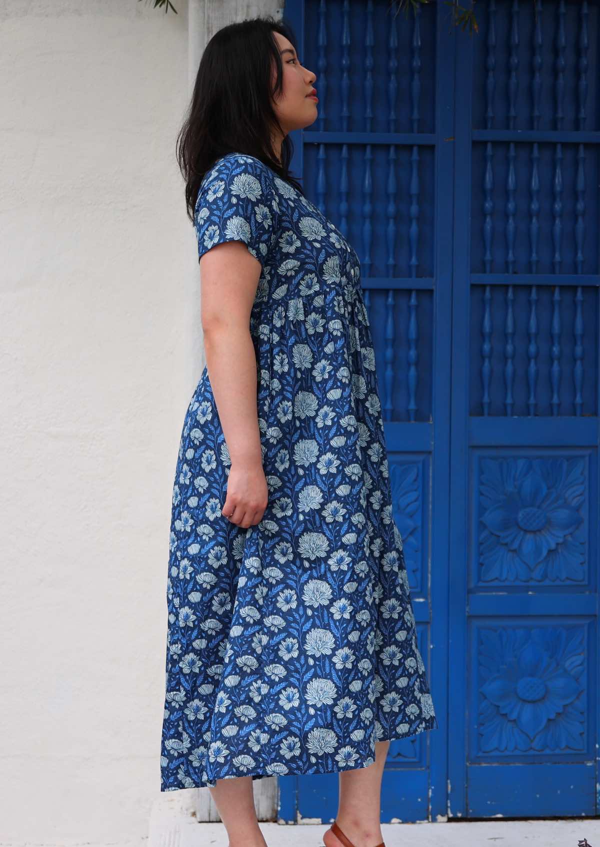 Model standing side on in front of blue door wearing 100 percent cotton navy blue button through v-neck dress with pockets