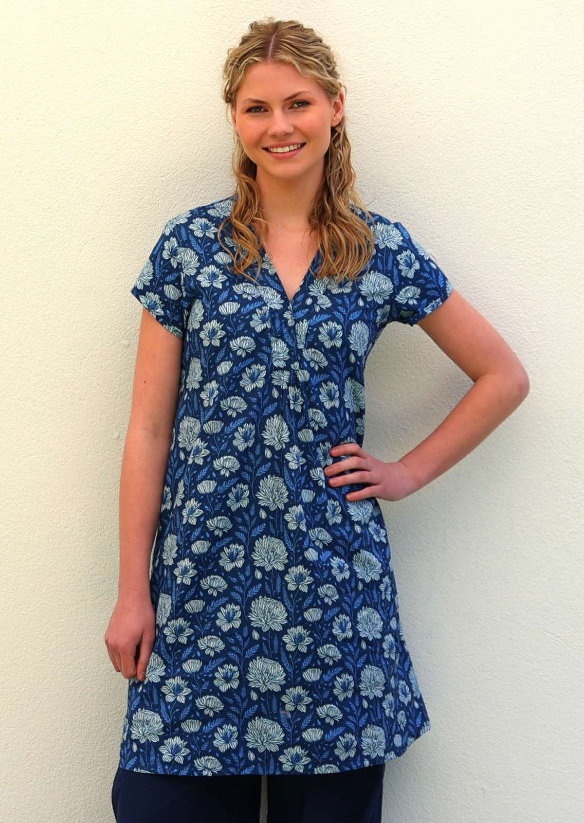 Woman with blonde hair wearing a blue cotton tunic dress with light blue and white floral print, in front of beige wall